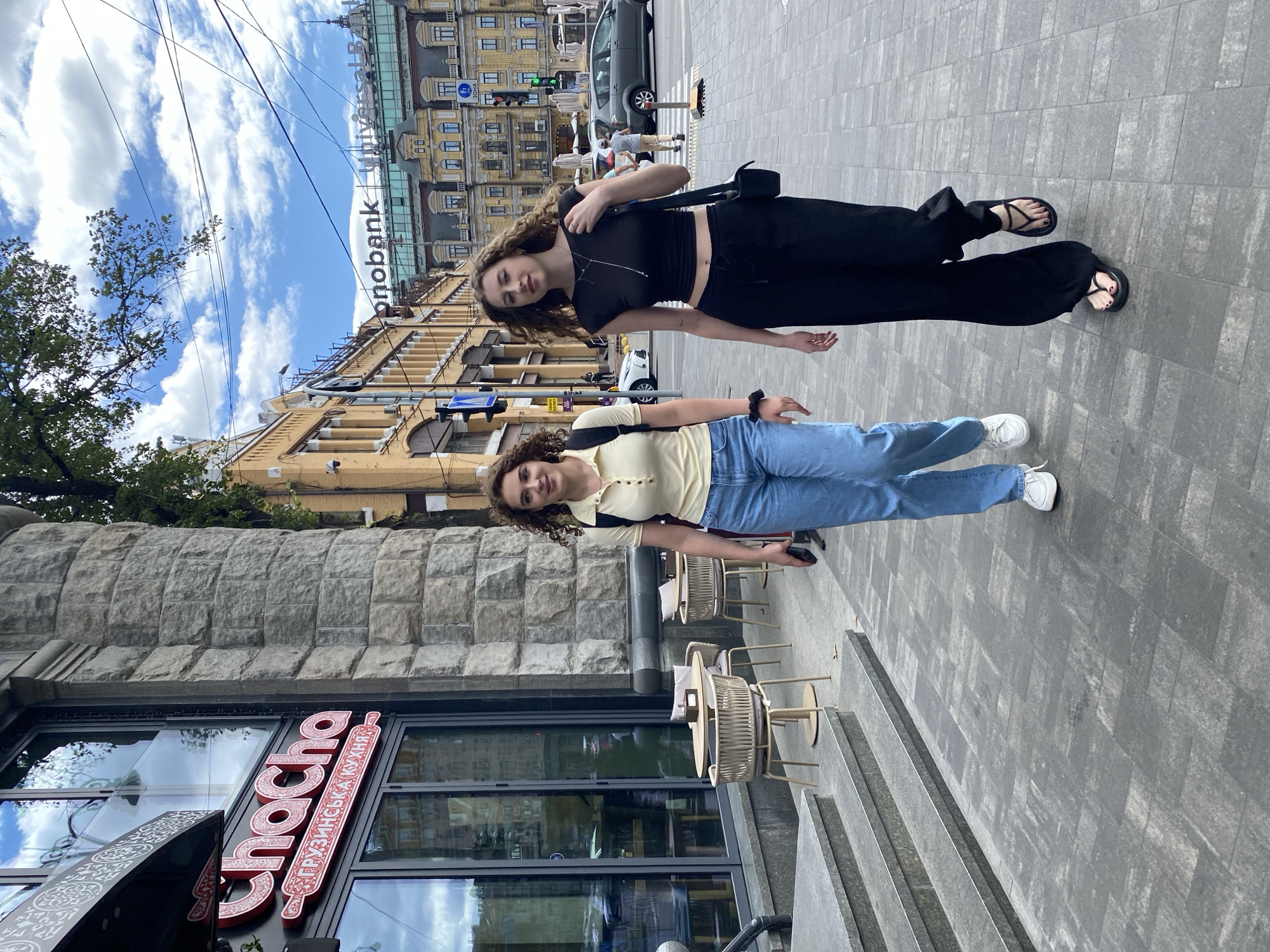 Two young women with curly brown hair walk on a paved city sidewalk. The woman on the left wears a yellow polo shirt, blue jeans, and white sneakers. She carries a black backpack. The woman on the right wears a black crop top, black flared pants, and black sandals. She carries a black shoulder bag. Both women are looking forward as they walk. Behind them, a restaurant with a sign that reads "Chacha" is visible. Further in the background, there are yellow buildings, a bank with the "monobank" logo, and a street with cars and traffic lights. The sky is blue with white clouds.