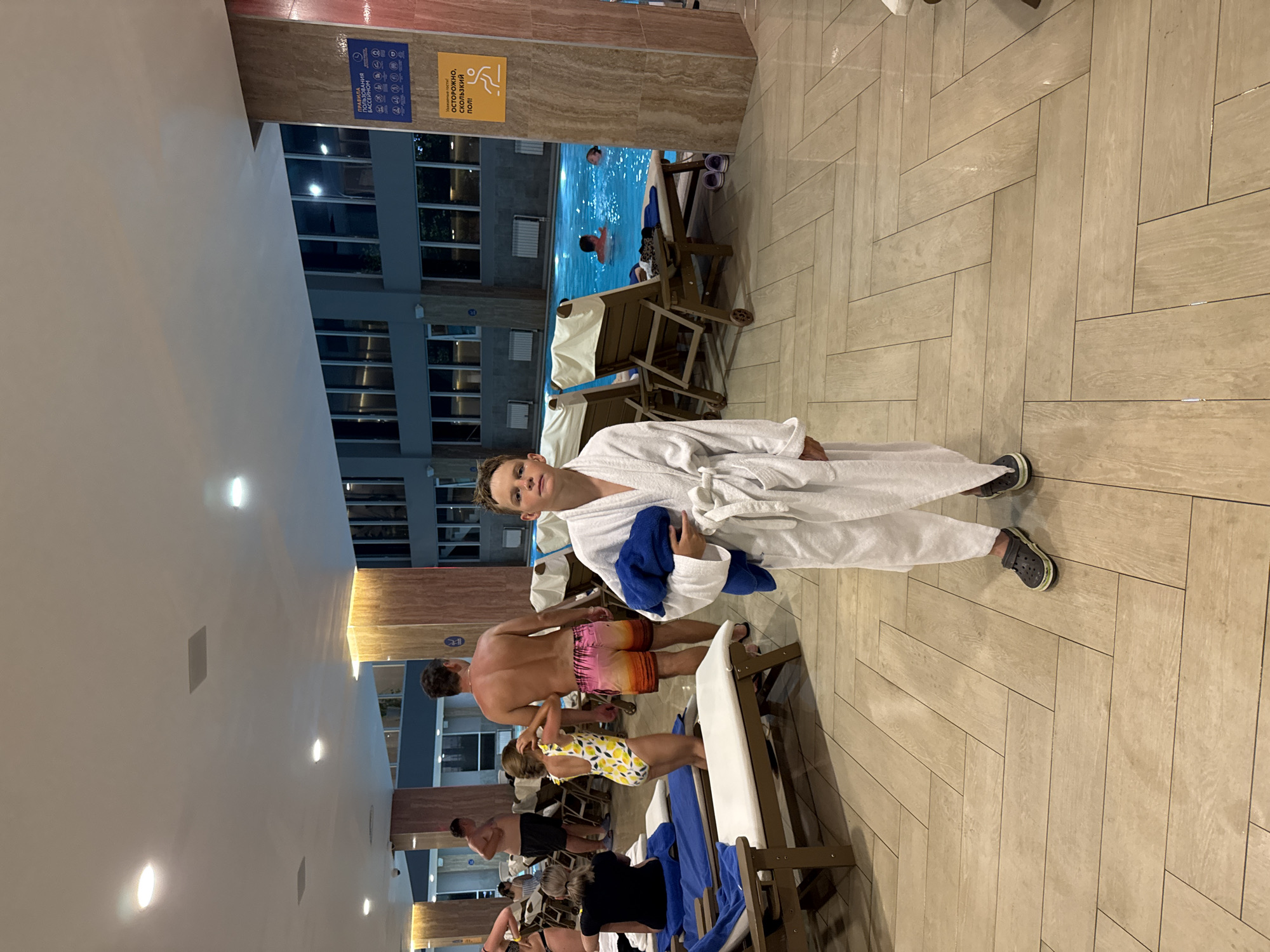 In a brightly lit indoor swimming pool area in Anapa, Russia, a young boy stands in the foreground wearing a white bathrobe and dark gray Croc-style shoes, holding a dark blue towel. He is looking directly at the camera with a neutral expression. Behind him, a swimming pool is visible with several people swimming. To the left of the pool, lounge chairs are arranged with towels on them. In the background, a tall man in pink patterned swim trunks is standing, and to his left, a young girl in a yellow and white lemon-print swimsuit is standing. Other individuals and poolside furniture are visible in the periphery. The floor is tiled with a light-colored wood-patterned laminate in a herringbone design. Two signs are mounted on a textured pillar on the right side of the image; one blue sign displays pictograms related to pool rules, and a yellow sign beneath it warns, "Attention guests, slippery floor." The lighting suggests it is evening or nighttime.