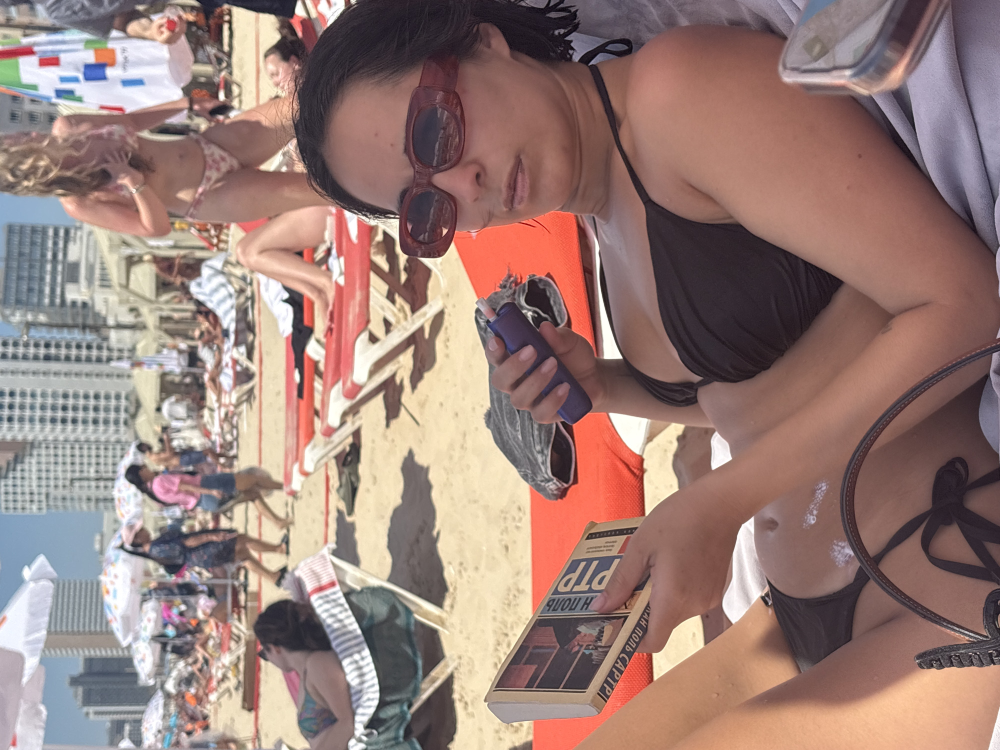 The image depicts a sunny day at a crowded beach in Tel Aviv, Israel. The foreground features a woman lying on a red beach chair, wearing a black bikini and large, dark sunglasses. She is holding a book in her left hand and what appears to be an electronic cigarette in her right hand, from which a wisp of vapor is visible. Sunscreen is applied to her stomach. In the background, many people are visible, some sunbathing on chairs, others walking on the sand, and several beach umbrellas providing shade. The architecture of Tel Aviv's skyline, characterized by numerous tall buildings, is visible in the distance, confirming the urban beach setting. The overall atmosphere suggests a typical recreational day at a popular city beach.