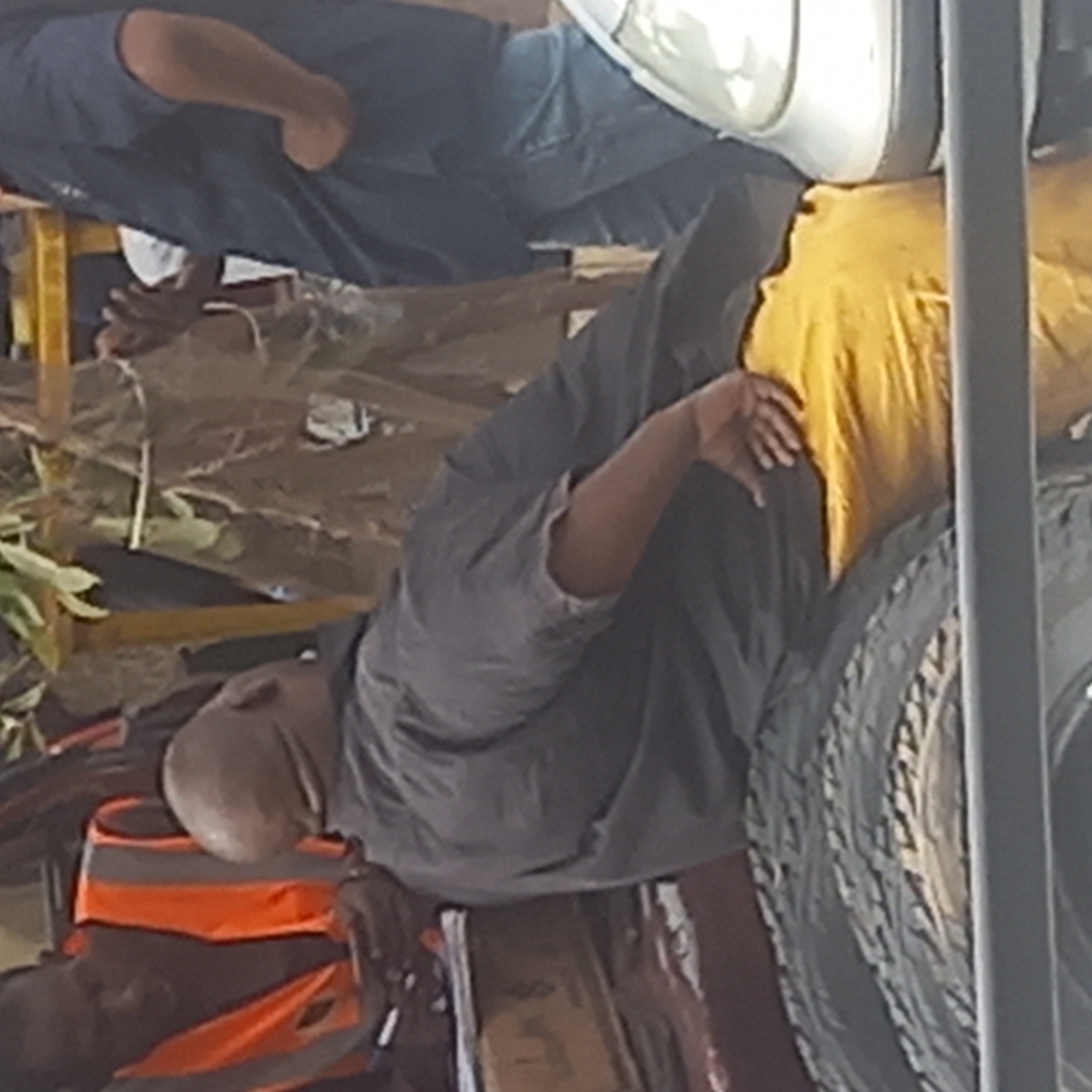 A street scene in Dadi Riba, Nigeria, shows several people involved in work. One person, wearing an orange safety vest, is seated and appears to be writing or taking notes. Another person wears a yellow cloth over their face while next to a stack of tires. Behind them, other people appear to be working at a makeshift table or work area. The scene has a cluttered and active atmosphere, typical of a working environment.