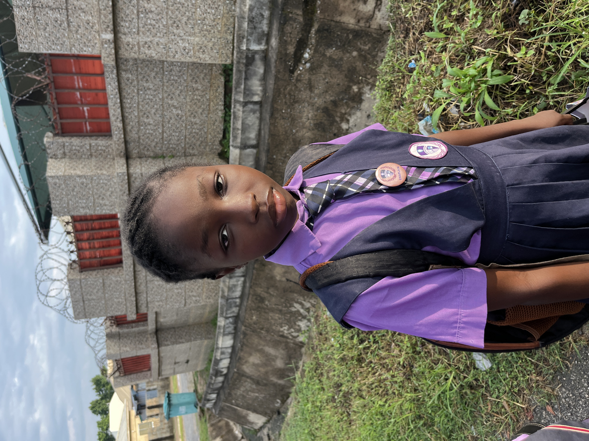 A young Black girl, approximately 8 years old, stands facing the camera outdoors in front of a building. She has dark skin, braided dark hair, and is wearing a school uniform. The uniform consists of a purple collared shirt, a dark blue jumper vest, a pleated dark blue skirt, and a patterned tie. A dark backpack is on her shoulders. She has two circular school badges pinned to her vest. One badge is light brown with the word "CHAPLAIN" and a crest. The other badge is pink and white with a crest. The building behind her is made of stone blocks with red-barred windows and topped with barbed wire. To the left of the building, a teal trash bin is visible next to a concrete structure. The ground is covered in grass and patchy dirt. The sky is overcast with some clouds. The setting appears to be a school or residential area in a city.