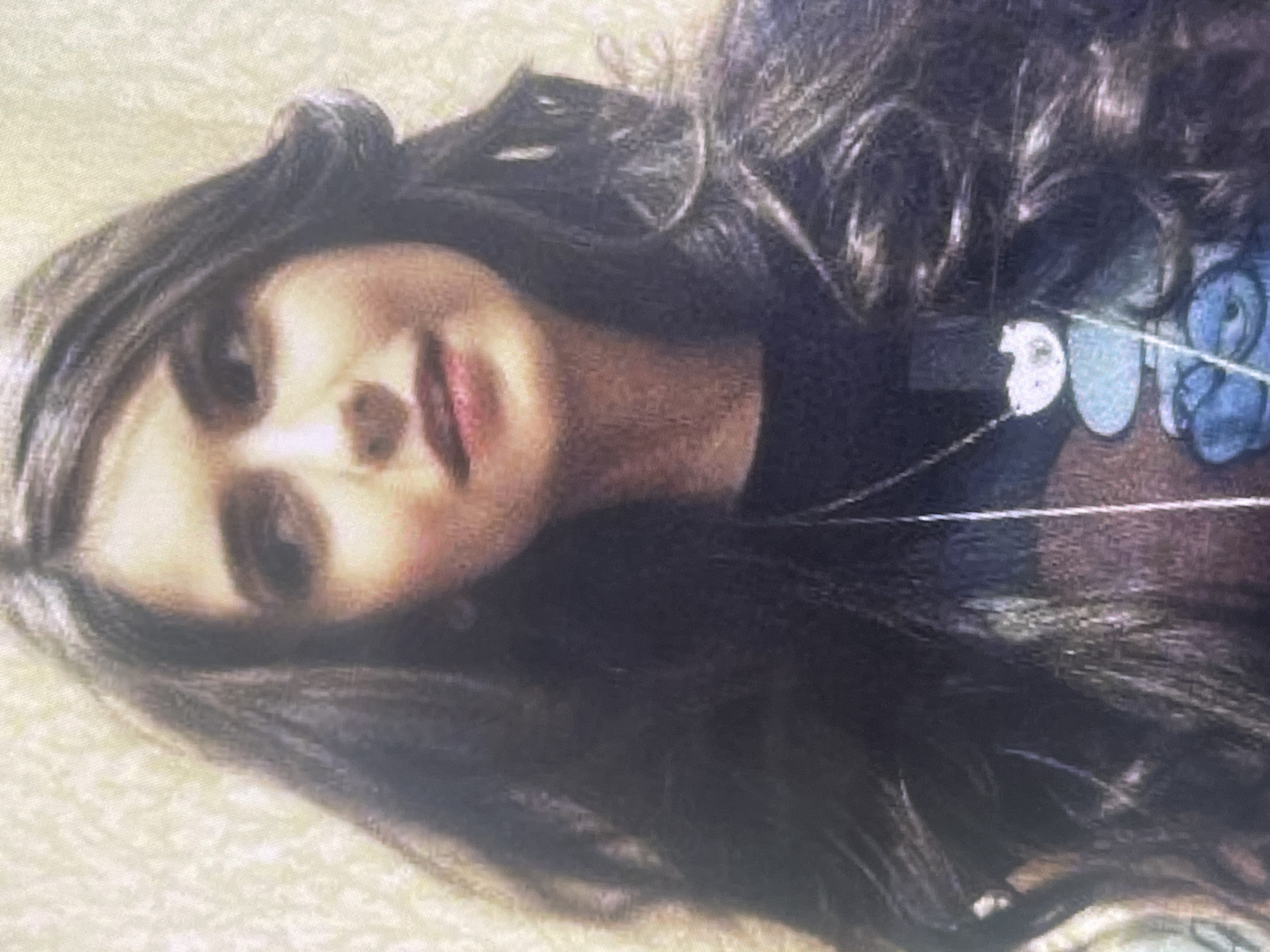 The image displays a young woman with dark, wavy hair, oriented sideways with her face gently turned towards the viewer. She has dark eyes and her lips are slightly parted, revealing a soft, natural pink color. Her long, dark hair cascades around her, framing her face with loose curls.
She is wearing a dark-colored top, possibly a t-shirt, with a light-colored design on the right side of her chest, featuring white and light blue elements that are not clearly identifiable as specific text or an emblem. A thin necklace with a small pendant is visible around her neck. The background consists of a light-colored, textured surface, which could be a blanket, pillow, or carpet, suggesting a relaxed indoor setting. The lighting is soft, casting a warm glow on her face, and the image has a slightly grainy quality. Her expression appears gentle or contemplative.
No specific activity is evident beyond her resting pose. There is no visible text. The location context provided is Poltava, Ukraine.