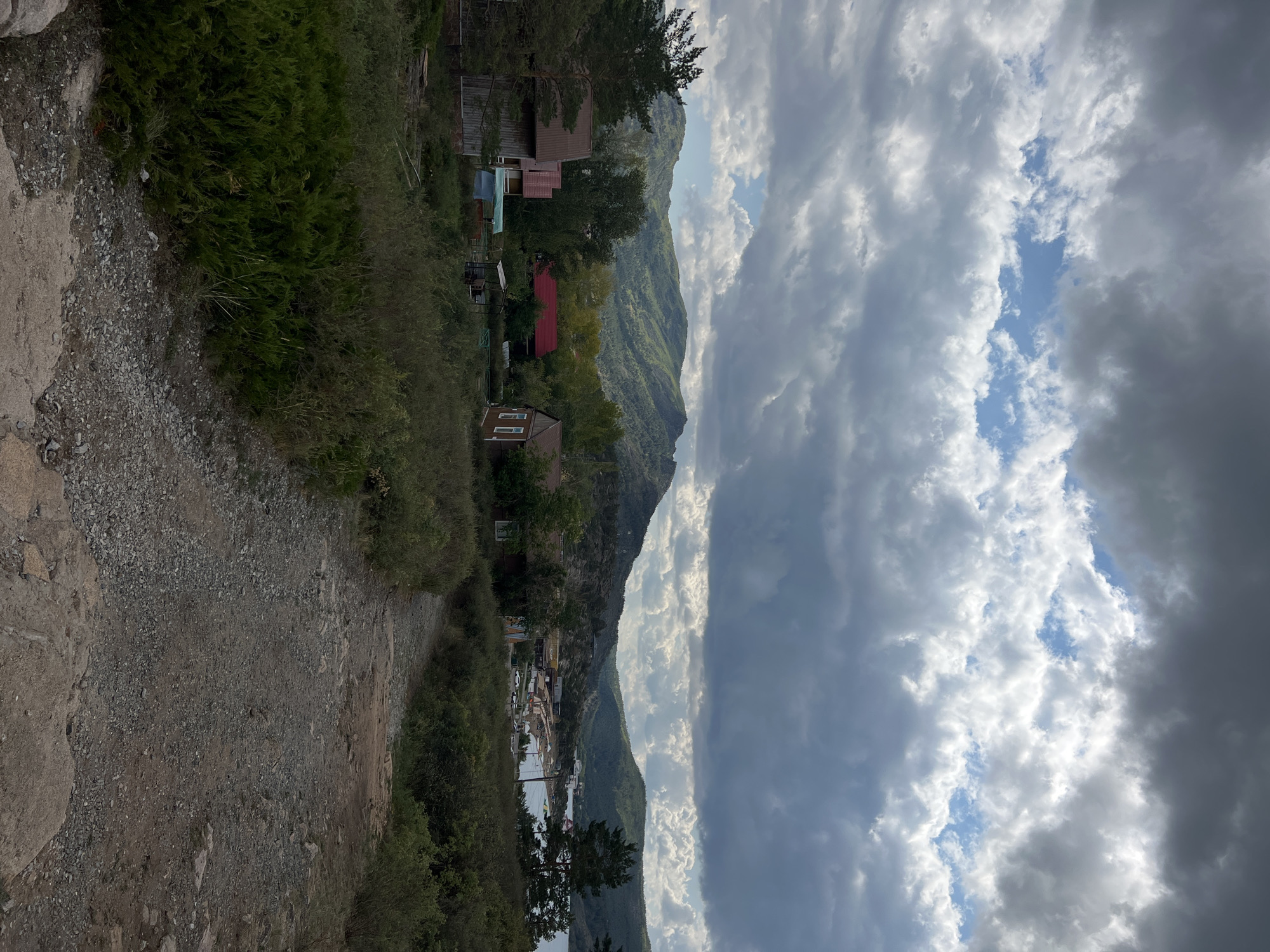 The image captures a rural landscape with rolling hills in the background and scattered houses in the mid-ground. A dirt path leads the viewer through overgrown vegetation and past several small wooden structures, some with red roofs. In the distance, a body of water is visible, bordered by more hills. The sky is filled with dramatic, cloudy formations, suggesting an overcast or possibly stormy day. The location is identified as Bayanaul, Bayanaul Rural District, Kazakhstan.