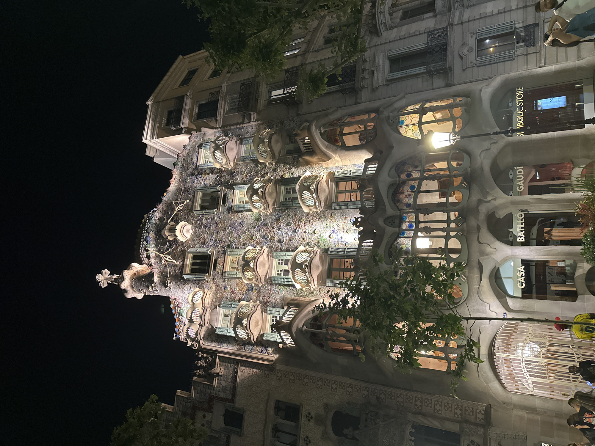 This is a nighttime photograph of Casa Batlló, a building in Barcelona, Spain designed by Antoni Gaudí. The facade of the building is illuminated. It features undulating stone work, colorful mosaics, and distinctive oval windows. Signs for "Casa Batlló", "Gaudí", and "Symbolic Store" are visible on the front of the building. Trees partially obscure the view of the building. People are present on the sidewalk below the building. The sky is dark.