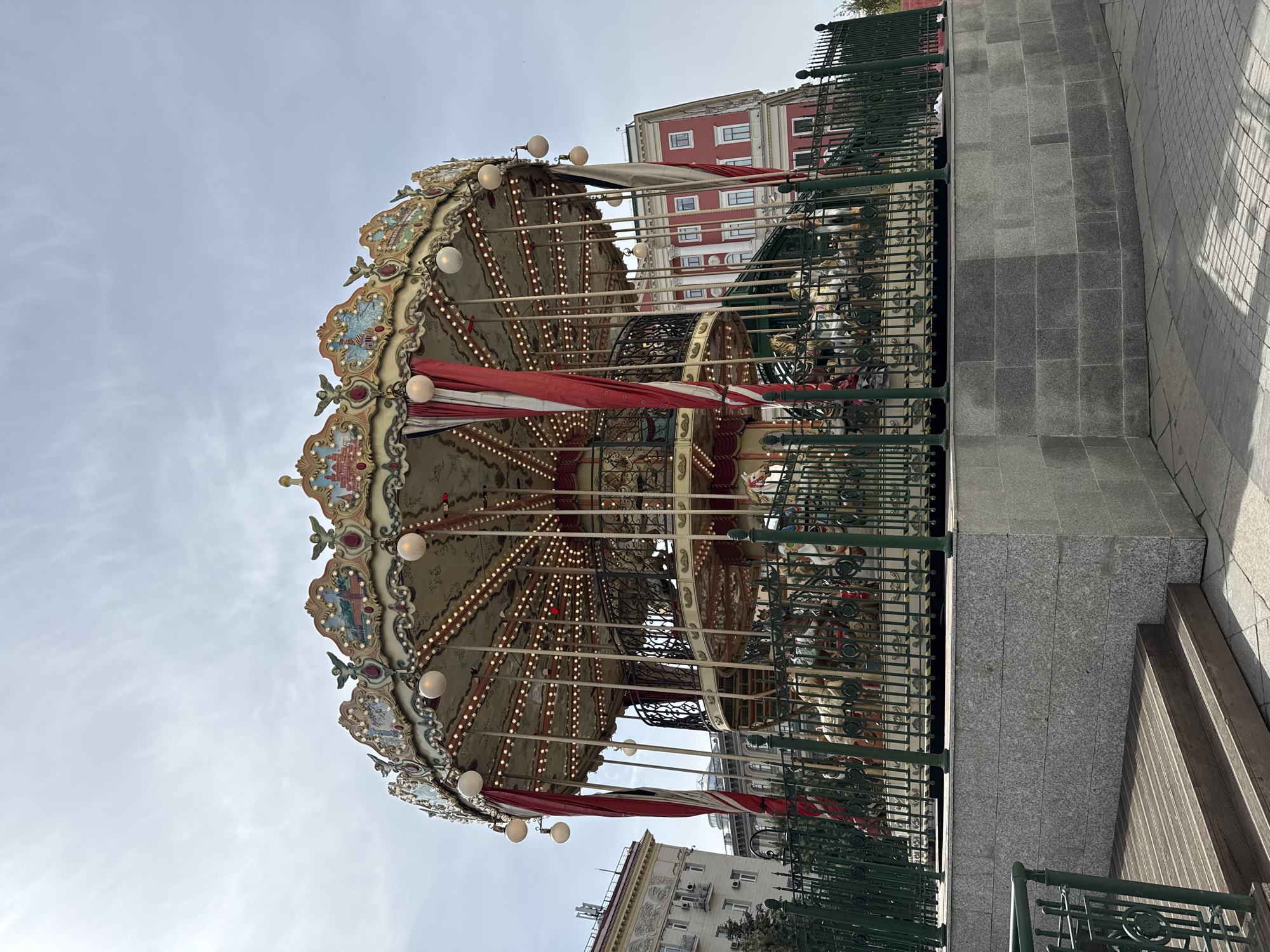 An ornate, traditional carousel stands as the central subject in an outdoor urban setting in Moscow, Russia. The carousel is elaborately decorated with multiple tiers featuring intricate carvings, decorative paintings in cream, blue, and red hues, and numerous small, glowing light bulbs that illuminate its structure. Red and white striped fabric awnings hang beneath its main canopy. Through the surrounding dark green, decorative metal fence, the classic figures of carousel animals, including horses, are visible inside.
The sky above is a uniform light grey, indicating an overcast day. Despite the daylight, the carousel's many lights are illuminated, contributing to its whimsical and inviting appearance. In the background, several buildings are present; one notably features a red facade with white window frames and architectural details, while another building is lighter in color. The area around the carousel is paved with grey, rectangular granite tiles. Shadows are visible on the paving, suggesting the presence of daylight. There are no people visible in the image, and the carousel appears to be stationary, presenting a calm and nostalgic scene in the city. No discernible text is visible on any of the objects or buildings.