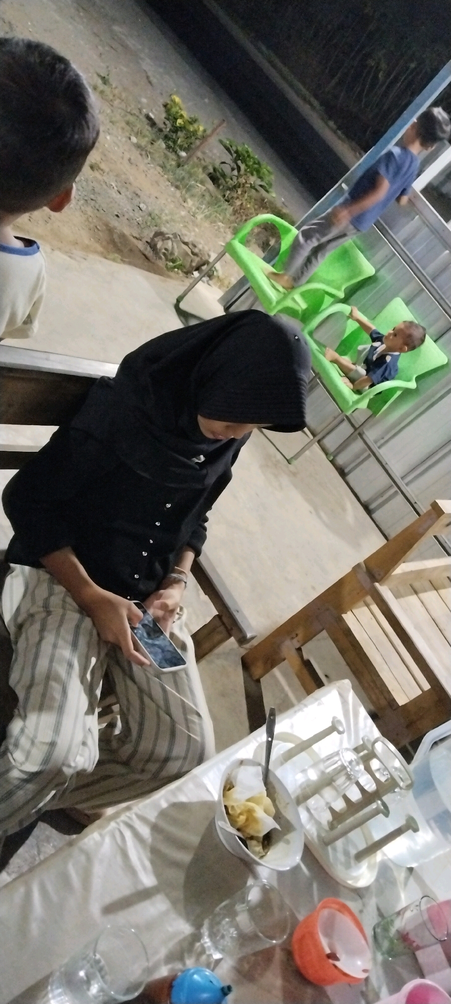 In an outdoor setting in Unknown, Indonesia, a group of people are gathered. A woman wearing a black hijab and striped pants is seated on a bench, holding a smartphone. She is looking down at the phone, seemingly engaged with it. To her right, a young boy in a blue shirt sits in a green plastic chair. Further to the right, another person, partially visible, is also seated in a green plastic chair. In the background, there are plants and a structure that appears to be made of metal and wood. In the foreground, a table is set with a white tablecloth, a bowl of food, several glasses, and a plastic container. The lighting suggests it is evening or night.