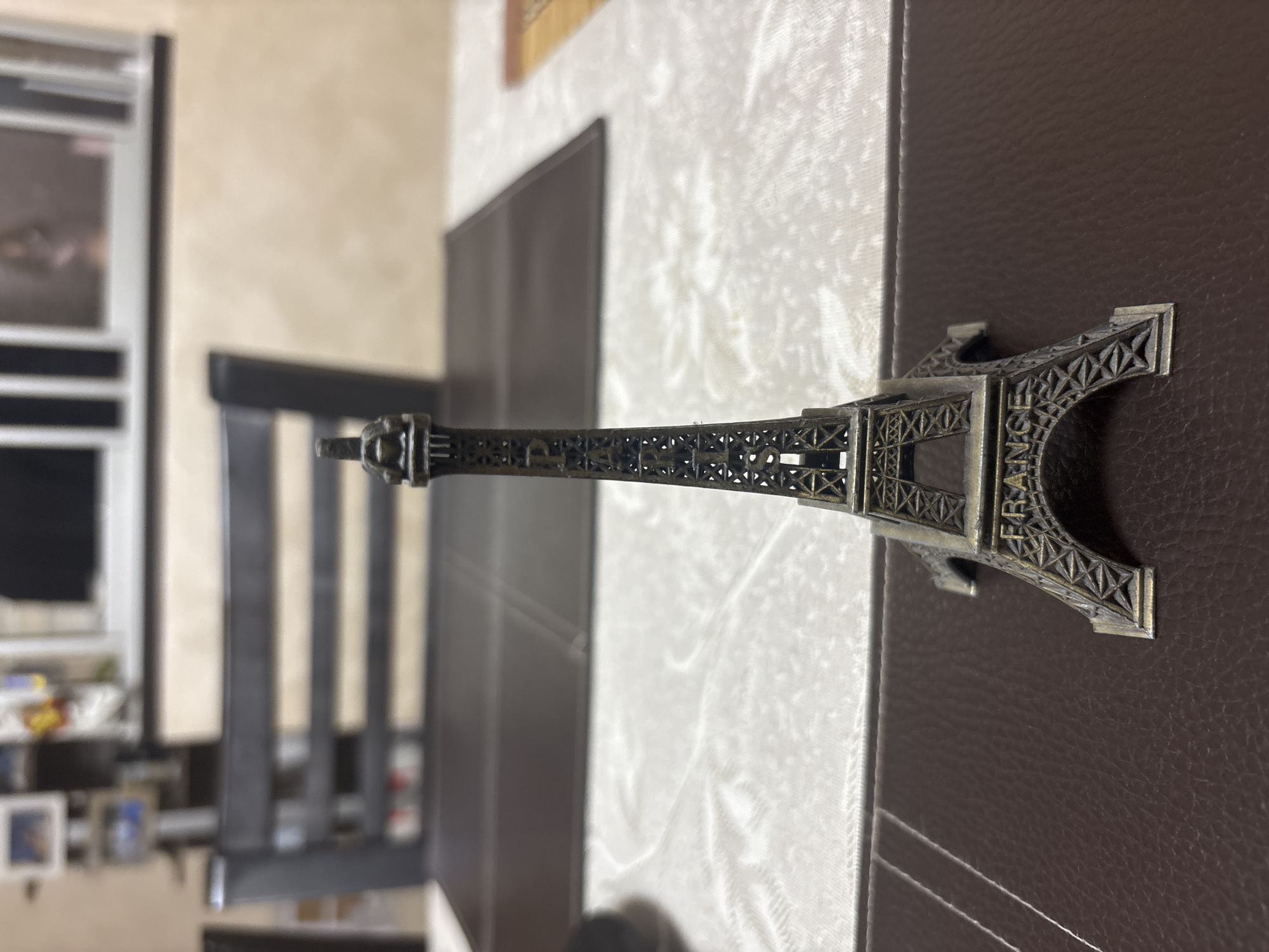 A sideways close-up shows a metal Eiffel Tower souvenir lying horizontally across two surfaces, a white fabric and a brown, textured surface. The tower is oriented from left to right, with the top of the tower on the left. "FRANCE" is etched on the bottom section. In the blurred background, a black chair is visible, along with a shelf with various items. There is no indication the photo was taken in Plovdiv, Bulgaria.