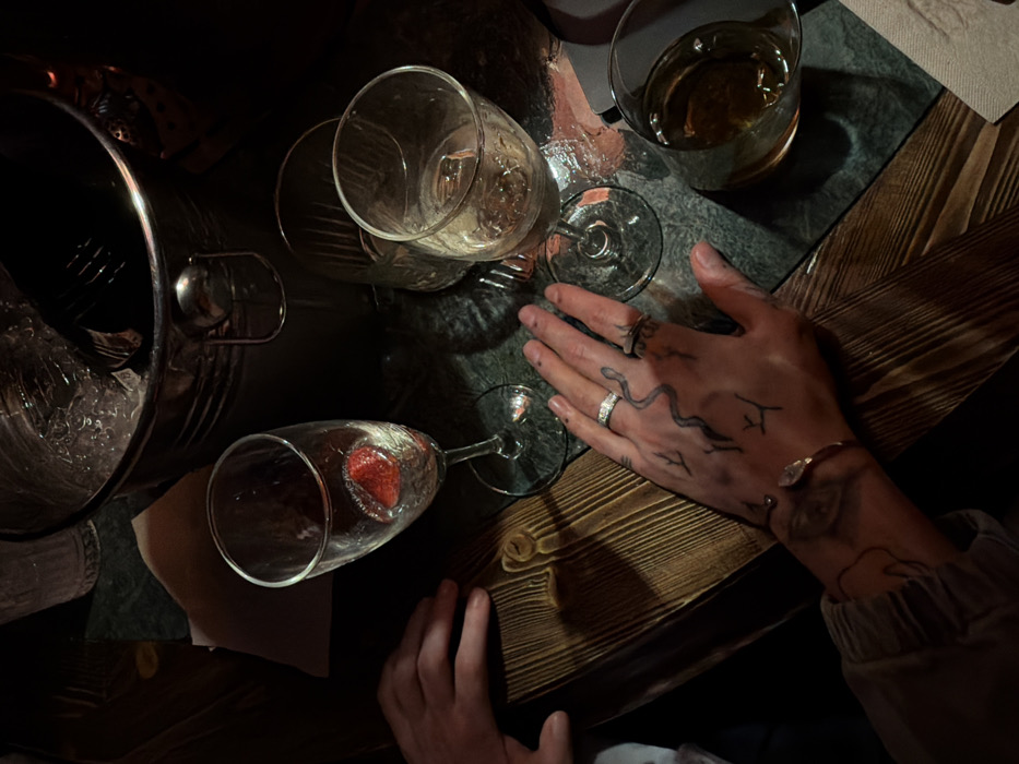 A dimly lit table setting features several empty wine glasses, one containing a strawberry. An ice bucket sits to the side. A person's hands are prominently displayed, adorned with rings and drawn snake tattoos. One hand rests near the glasses, while the other lies out of focus. The table is a mix of wood and dark stone or tile, adding to the intimate, possibly post-celebratory atmosphere.