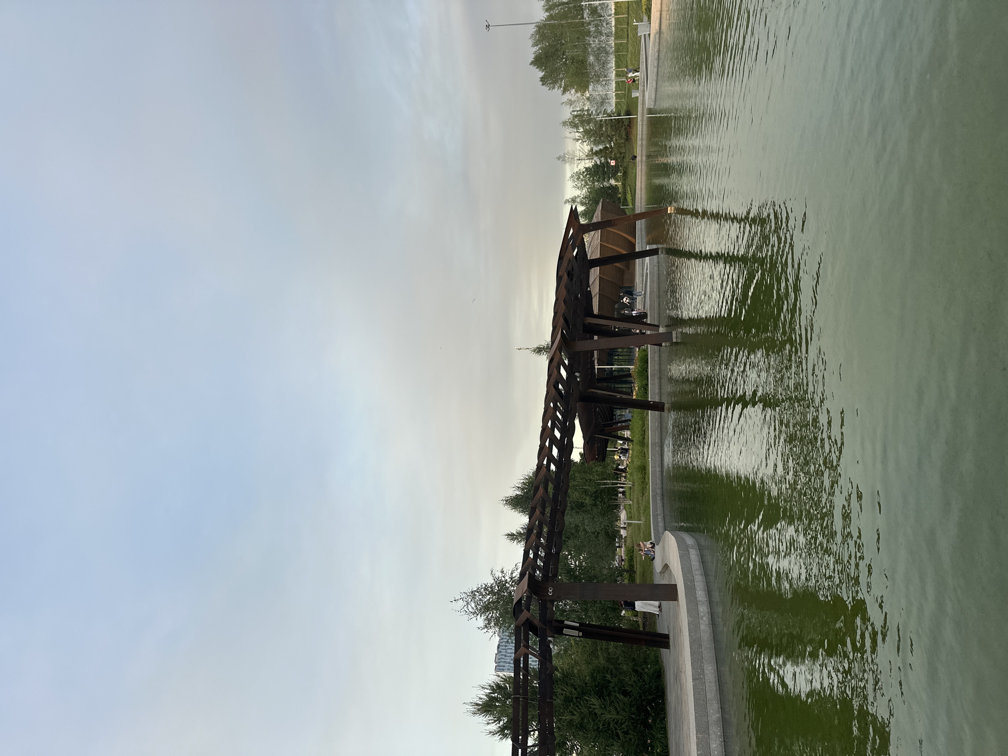 The image shows a view of a park with a pond and a modern wooden structure in the city of Moscow, Russia. The pond's water is a murky green and reflects the sky and the surrounding structures. To the left, a curved concrete embankment borders the pond, with people seen walking and sitting on it. A tall, dark wooden pergola-like structure with a lattice roof extends over the water and onto the embankment, supported by thick wooden pillars. Behind the pergola, there are lush green trees, some appearing to be evergreen, and in the distance, a modern building with a distinct architectural design is visible. The sky is overcast with soft, diffused light, suggesting it might be early morning or late afternoon. To the right, the park continues with more trees and manicured lawns, and a white fence can be seen in the far distance. Several people are scattered throughout the park, engaged in various activities such as walking, sitting, and standing. The overall atmosphere is serene and public, indicative of a well-maintained urban park designed for relaxation and recreation.