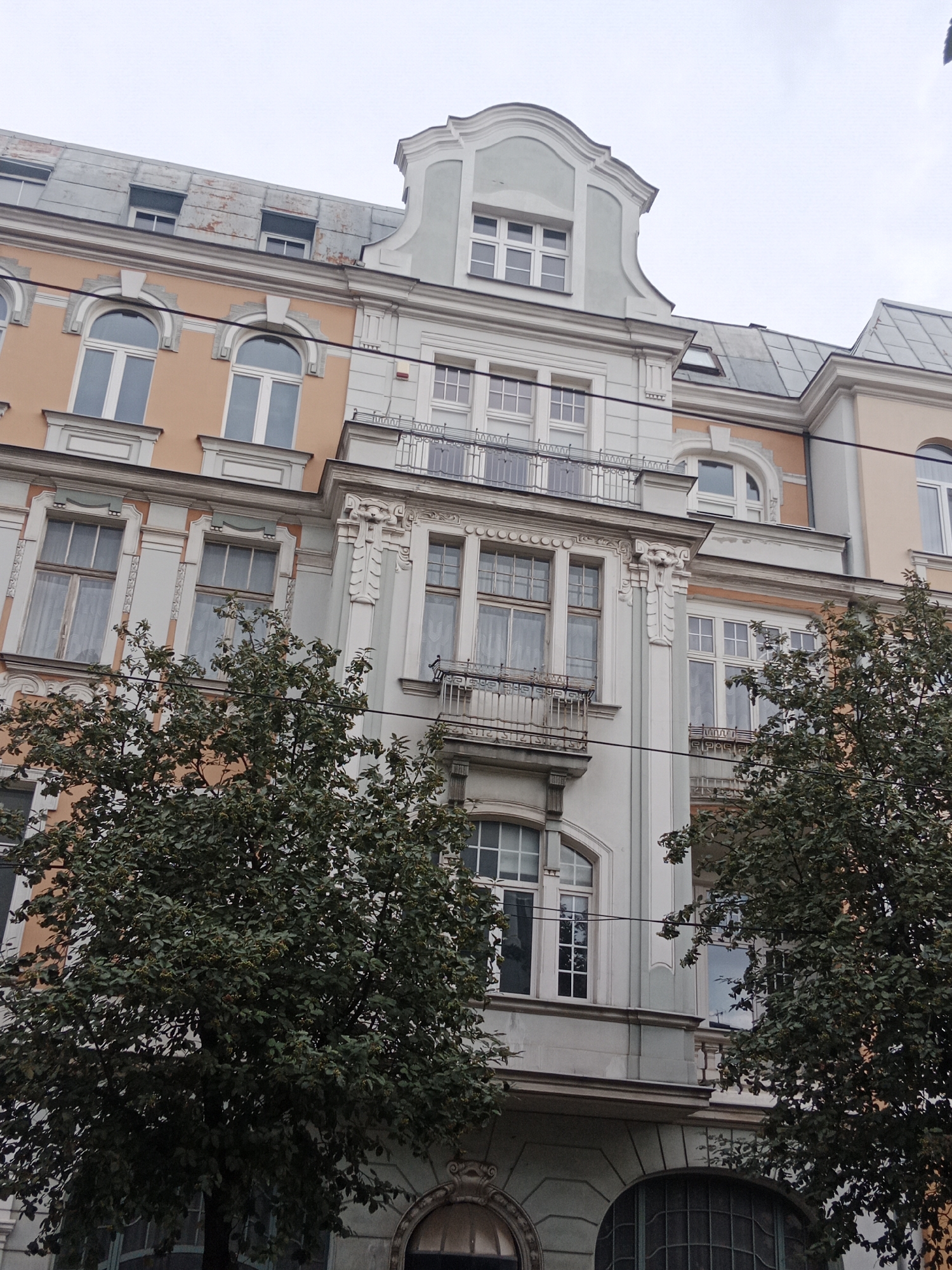 This is an eye-level, exterior, medium shot of the facade of an old European apartment building, likely in the city of Bydgoszcz, Poland. The building features several stories with a mix of architectural styles, including ornate decorative elements around the windows and balconies. The building's exterior is painted in muted colors of peach, light blue, and grey. Several large trees with dark green foliage are situated in front of the building, partially obscuring the lower floors. The sky is overcast and grey. There are no people visible in the image. The scene appears to be a daytime shot.