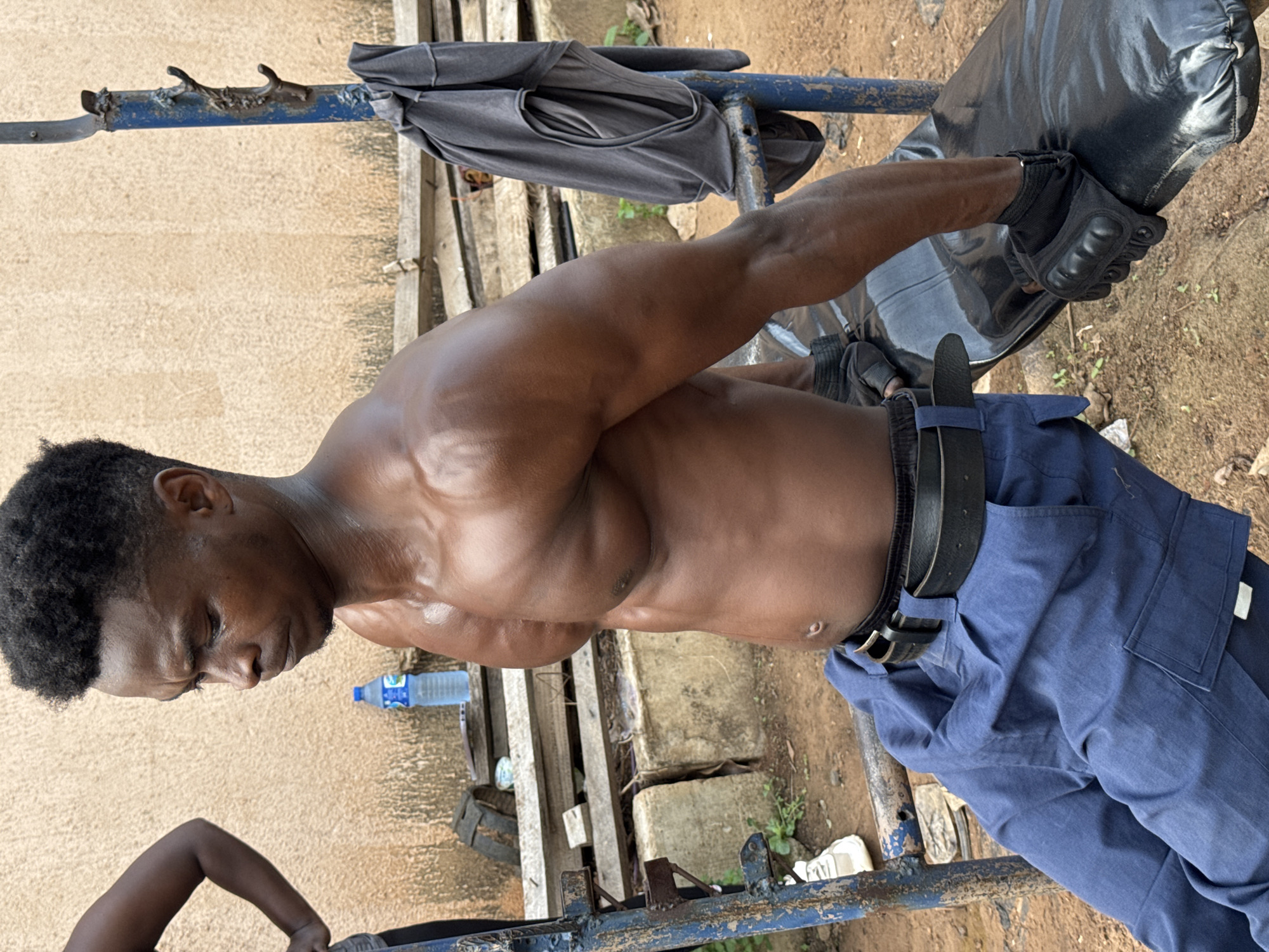 An undated, outdoor, medium shot captured in Akwanga, Nigeria, shows a shirtless Black man with dark skin, short, curly black hair, and a muscular physique, wearing blue cargo pants and a black belt, as he sits on a weight bench. He is wearing black weightlifting gloves on both hands, and his left hand is resting on the bench. His right arm is extended and resting on his right thigh. The man appears to be looking down and concentrating.
The background is somewhat blurry, but reveals a rough, textured tan wall, a blue metal workout structure, a gray t-shirt hanging on the structure, a plastic water bottle on a wooden surface, wooden planks, concrete blocks, and dirt. There are also hints of green foliage. The overall impression is a gritty, outdoor gym environment.