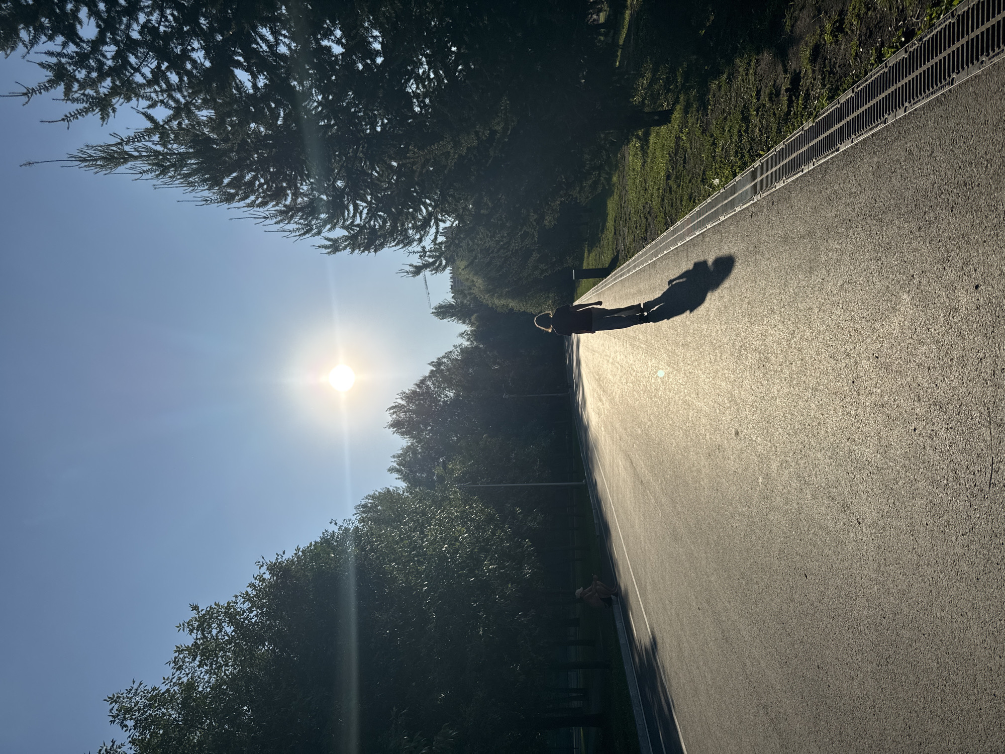 In Ufa, Russia, a person is captured in silhouette walking on a paved path under a bright, low-hanging sun. The sun creates a lens flare effect and casts a long shadow of the person onto the path. The path is bordered by a grassy area and tall coniferous trees on the right, and a line of deciduous trees on the left, casting dappled shade. Another person is seated on the curb to the left, appearing to observe something on the ground. The sky is clear and blue, indicating a sunny day. The scene is set in a park or a similar outdoor recreational area.