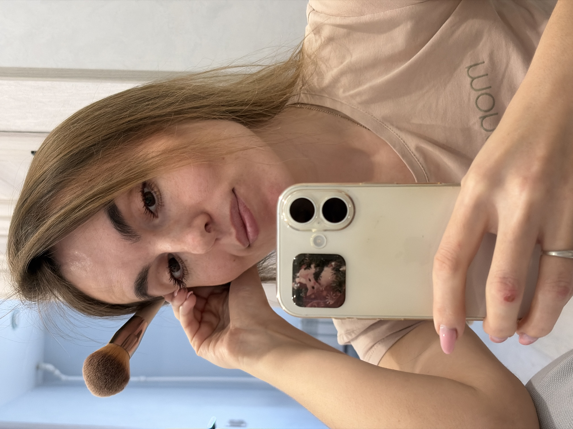 A young woman is taking a selfie. She's holding a phone with a dual-lens camera and a floral design on the back. In her other hand, she holds a makeup brush near her face. The woman has light brown hair, dark eyebrows, and pink lipstick. She is wearing a light pink t-shirt with text on it. A ring is visible on her finger. The photo appears to be taken indoors, and the background includes a door frame and a pale blue wall. The image does not contain details to confirm a location in Sarapul, Russia.