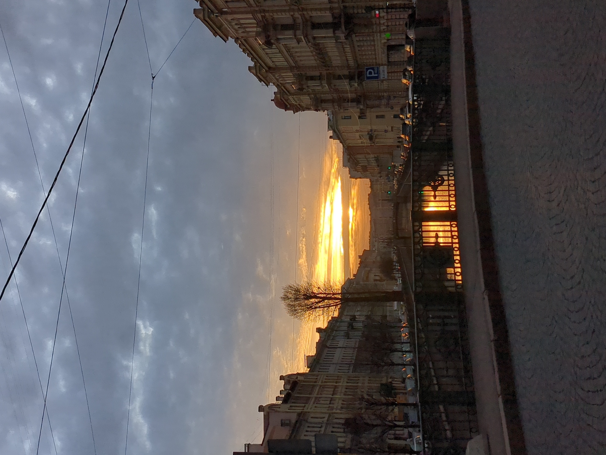 This image captures a sunset view along a canal in Saint Petersburg, Russia. The sky is a mix of grey clouds and bright orange streaks where the sun is setting. Buildings line both sides of the canal, exhibiting classic European architecture. A bare tree stands silhouetted against the sunset. Reflections of the sunset and building lights shimmer on the water's surface. Power lines are visible against the sky on the left side of the image. The edge of a sidewalk is visible in the foreground on the right.