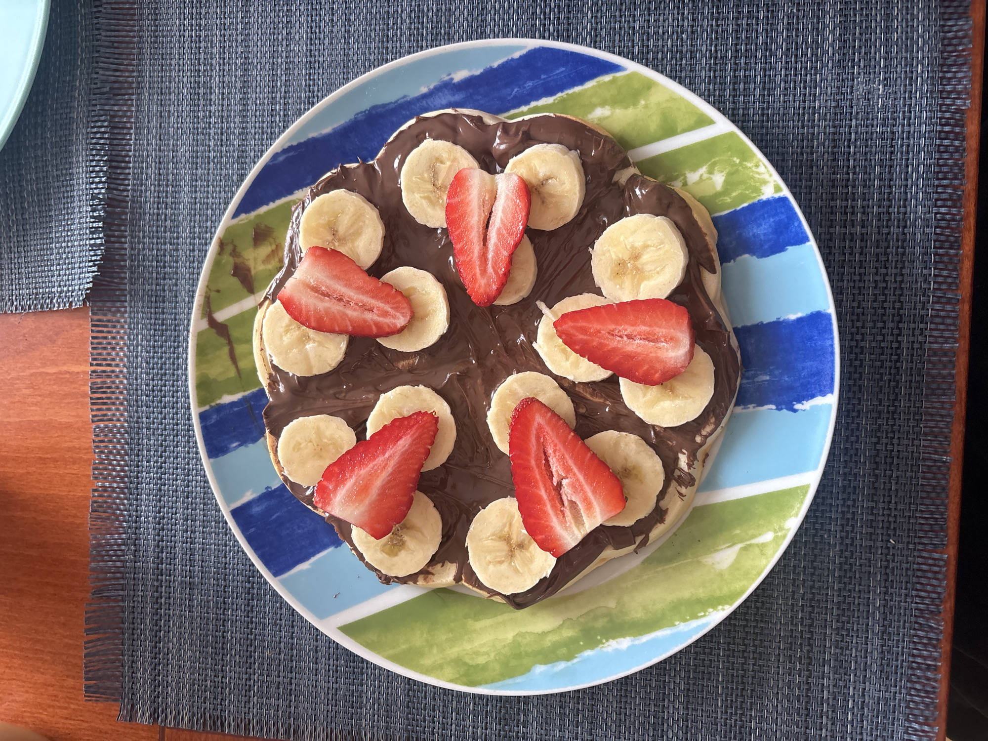 A top-down, close-up shot depicts a colorful plate of dessert placed on a dark blue woven placemat. The plate features alternating stripes of blue, light blue, green, and white. The dessert consists of a base covered in chocolate spread, topped with sliced bananas and strawberries. The strawberries are halved and arranged strategically around the plate. The placemat has a woven texture. The left edge of a light blue plate is visible to the left of the main plate. The scene appears to be indoors, possibly in a home or cafe. The location is not discernible within Sofia, Bulgaria based on the given image.