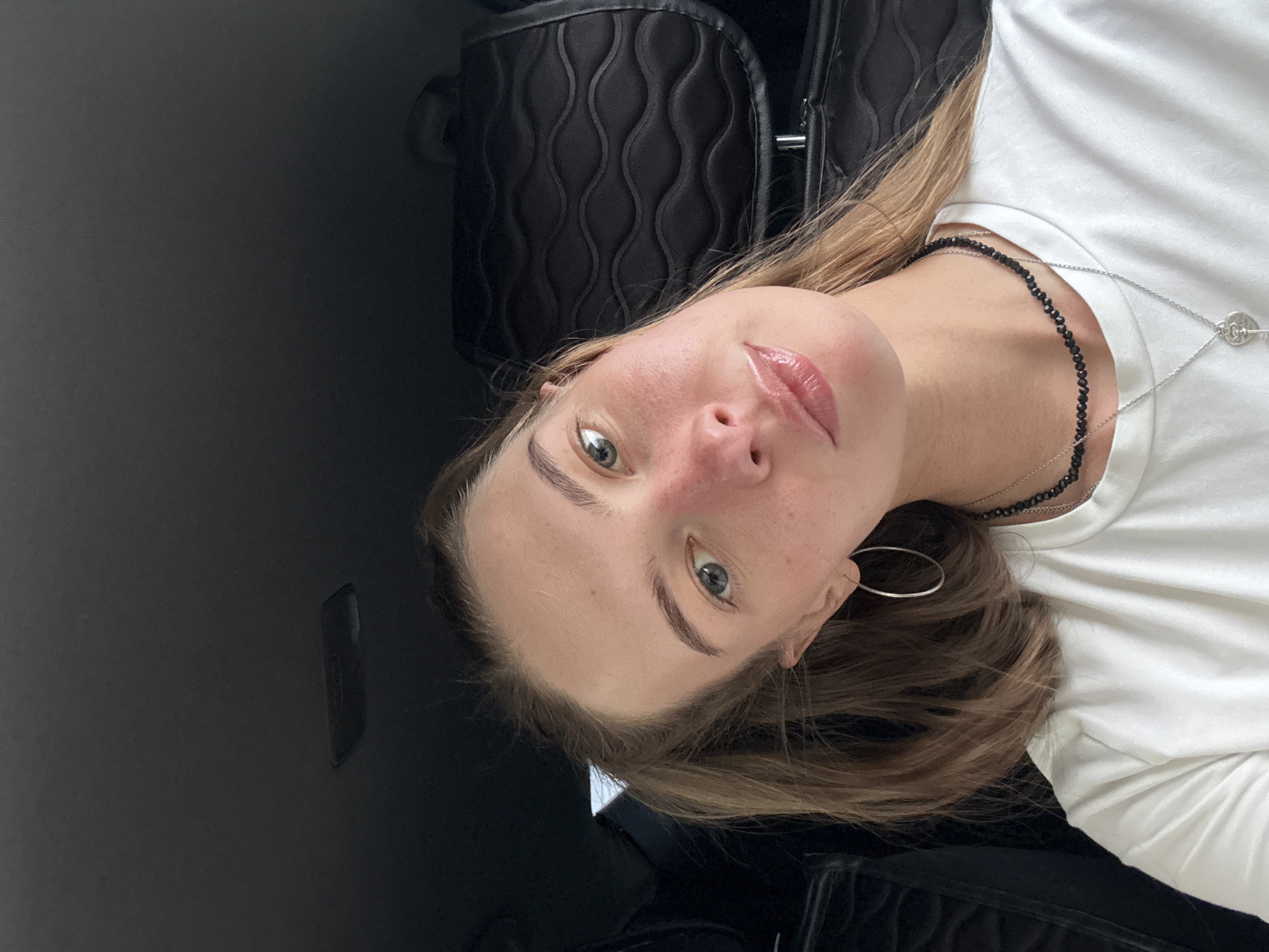 The image is a selfie taken by a young Caucasian woman inside a vehicle. She is wearing a white t-shirt and several necklaces, including a black beaded one and a silver one with a key pendant. She has on large hoop earrings. Her hair is pulled back into a ponytail, and she has light skin with freckles and rosy cheeks. She is looking directly at the camera with her head tilted. The background shows the interior of a car, with black leather seats and the ceiling. There is no information within the image to suggest the location is in the city of Perm, Russia.
