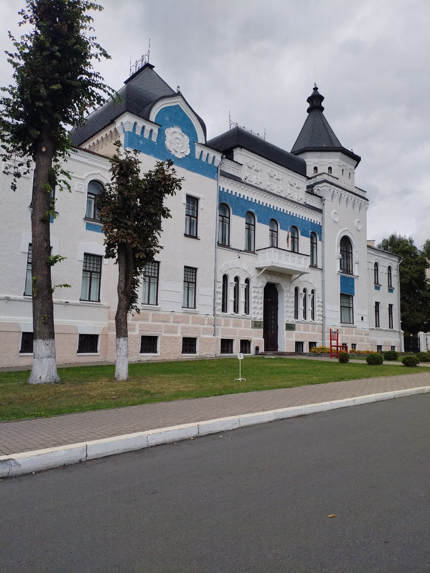 The image shows the facade of a historic building located in Mahilyow, Belarus. The building is two stories high with a white exterior, accented with blue decorative elements along the roofline and around some windows. It features a prominent tower with a dark, conical roof and a pointed spire. Multiple windows are visible, some rectangular and others arched. Two trees stand in front of the building, their trunks whitewashed near the base. A paved sidewalk runs along the front of the building, separated from a grassy lawn by a curb. The foreground shows an asphalt road. The sky is overcast, suggesting cloudy weather. There are no people visible in the image.