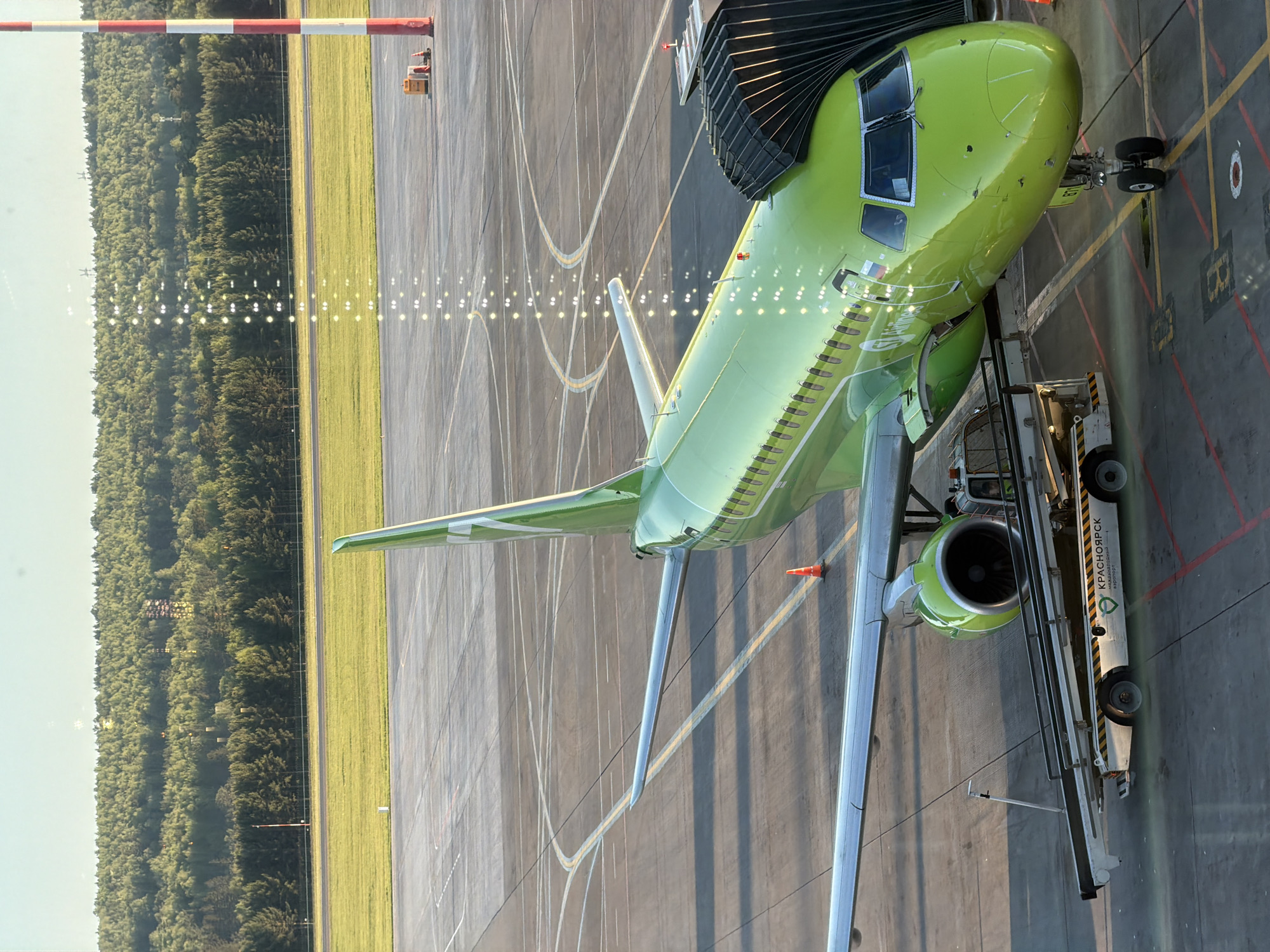 The image shows a green S7 Airlines Airbus A320neo aircraft parked at a gate at the Емельяново Airport (KJA) in Krasnoyarsk, Russia. The aircraft is connected to a jet bridge. A ground support vehicle is parked alongside the aircraft. A runway with lights and a grassy area bordered by trees are visible in the background.
