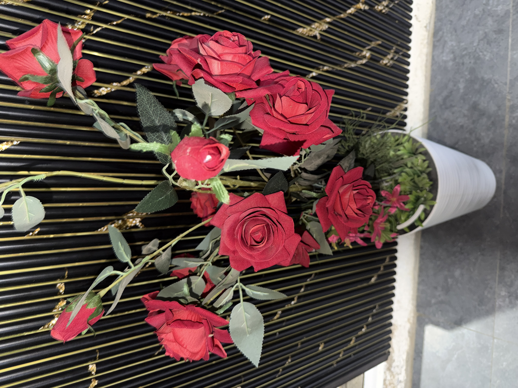 This media file features a close-up, overhead view of a bouquet of artificial red roses arranged in a white cylindrical vase. The vase is placed on a surface covered with black horizontal blinds that have thin gold lines running through them. The background transitions from the blinds to a mottled grey surface, suggesting an indoor setting possibly near a wall. The lighting is natural and appears to be from above, casting soft shadows. The roses are in various stages of bloom, with some fully open and others still budding. The leaves are a muted green, and the stems are visible, wrapped in green material. The overall impression is one of artificial elegance, with the bright red of the roses contrasting with the dark blinds and white vase. There is no visible text in the media file. The setting does not contain any identifiable landmarks or cultural elements specific to Khumsan, Uzbekistan, other than the provided context.