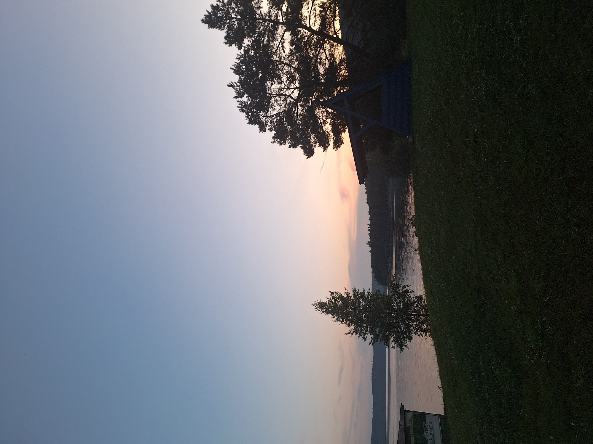 The image depicts a serene evening landscape with a clear sky transitioning from light blue to soft orange hues near the horizon. A calm body of water, likely a lake or river, stretches across the mid-ground, bordered by a tree-lined shore in the distance. In the foreground, a neatly mowed grassy hill slopes downwards towards the water. A single, slender tree stands prominently on the left side of the hill, while on the right, a dark silhouette of pine trees frames a triangular wooden structure, possibly a gazebo or shelter, illuminated by dim light. A small, weathered building with visible signage is partially visible on the far left, near the water's edge. The overall scene suggests a tranquil, natural setting at dusk, potentially in the vicinity of Атамановка, Russia, given the context provided.