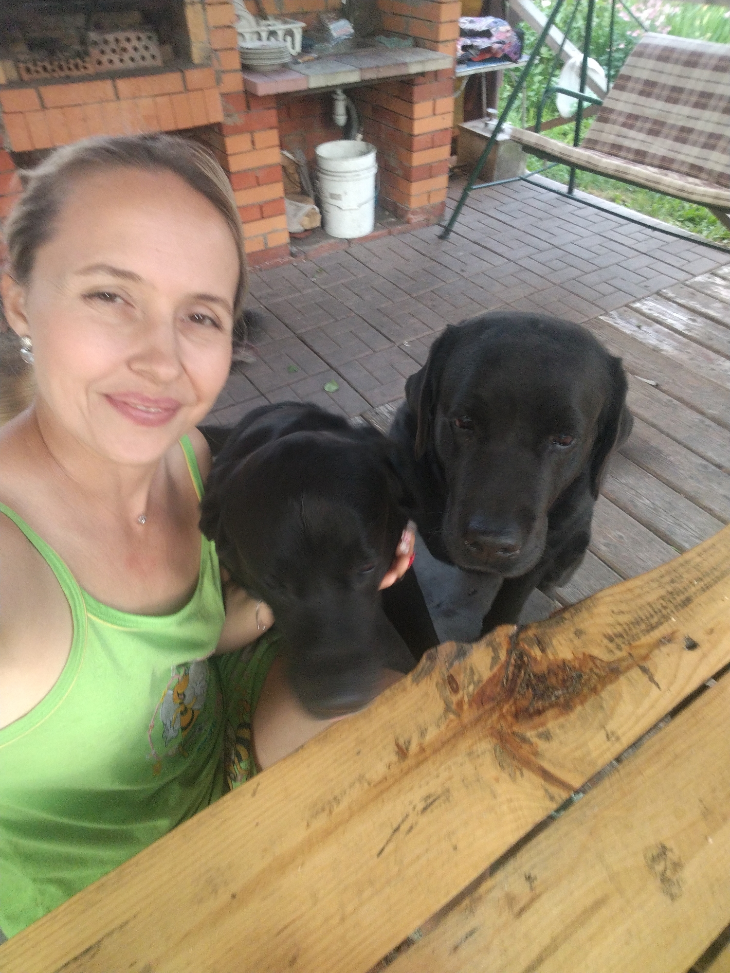 A woman with blonde hair pulled back and wearing a green tank top is taking a selfie with two black Labrador retrievers in an outdoor setting. The woman is smiling and has a small diamond pendant on a chain around her neck. One dog's head is close to the woman's face, and the other dog is looking directly at the camera. They are on a wooden deck in front of a brick structure with a grill. In the background, a swing set with a striped cushion and greenery can be seen. The overall scene appears to be a backyard or patio area. The specific location in the city of Рассвет, Russia cannot be determined from the image alone.