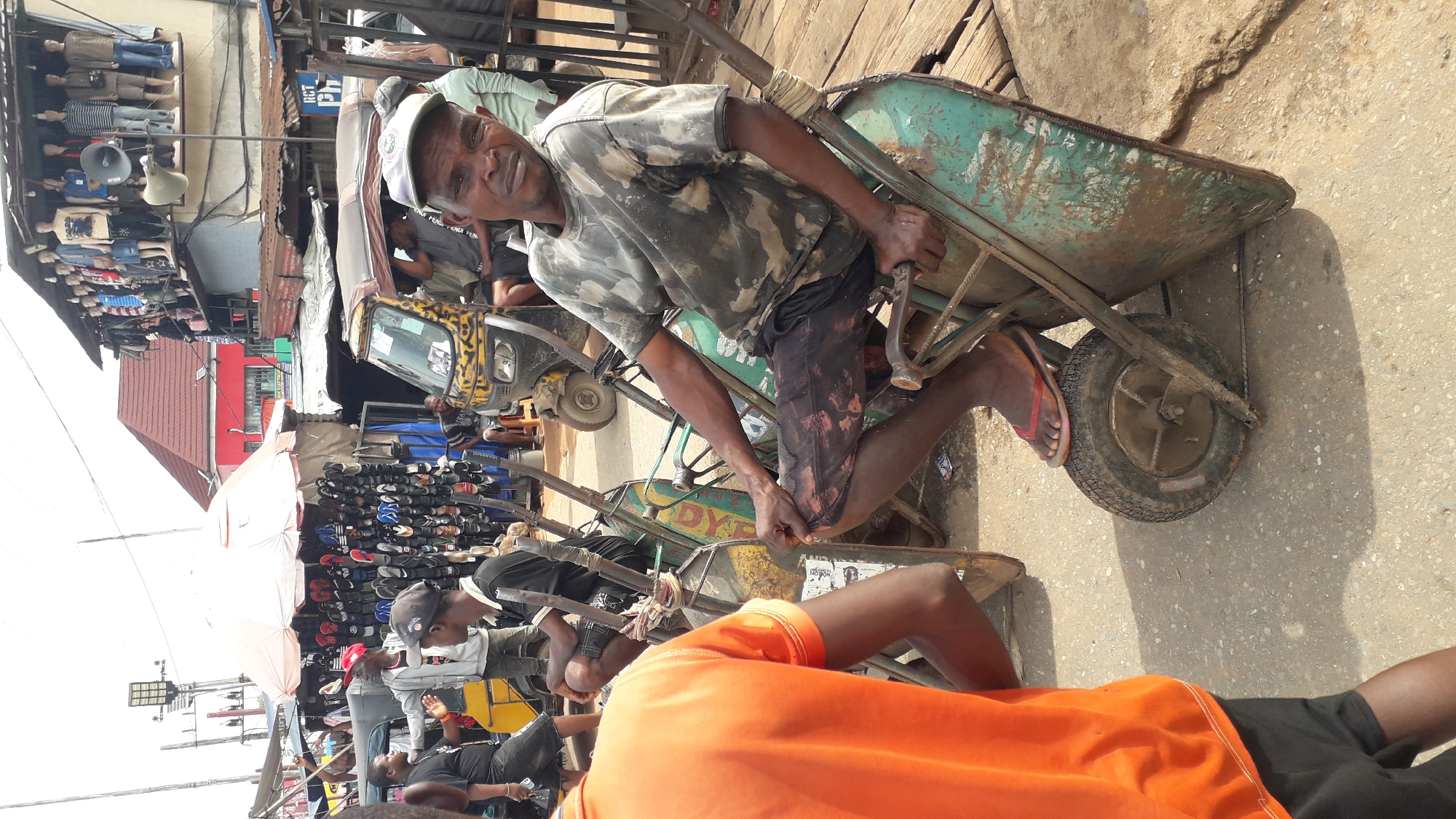 This is a medium shot, eye-level photograph taken in Onitsha, Nigeria, likely within the bustling Bridge Head market area. The scene is dominated by an outdoor marketplace environment. In the foreground, a man wearing a camouflage t-shirt and dark shorts with red paint stains sits on a teal-colored wheelbarrow. He is looking directly at the camera with a neutral expression. His left hand rests on the wheelbarrow's handle, and his right hand is placed on the wheelbarrow's bed. A second wheelbarrow is positioned to his left.
Behind the man, a yellow motorized three-wheeled vehicle, commonly known as a Keke Napep, is parked, with several people visible inside and around it. To the left of the Keke, a stall draped with a large white umbrella displays an abundance of footwear, primarily flip-flops, hung neatly. Further in the background, on an elevated platform or building, mannequins dressed in various clothing are displayed. Red-roofed buildings and stalls with corrugated metal roofs are also visible, indicative of the dense commercial activity typical of Onitsha markets. The ground is paved with asphalt. The overall impression is one of a vibrant and crowded marketplace.