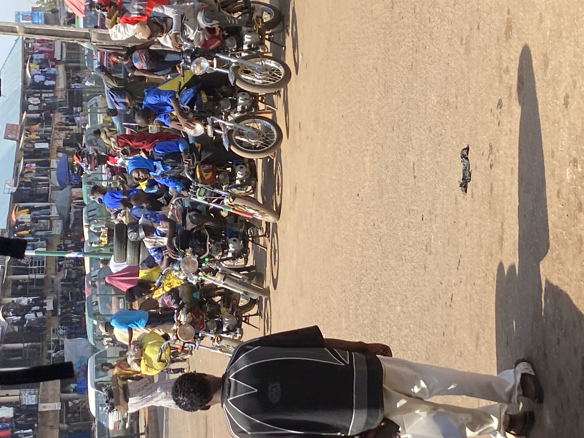 The image depicts a busy street scene in Bukuru, Nigeria, densely populated with people and motorcycles. A row of motorcycle taxis line the left side of the street, their drivers present and possibly soliciting fares. Buildings on the left house shops, filled with merchandise. A man in a black jacket and white pants stands in the foreground, partially obscuring the scene. The road surface is bare except for a piece of debris and the shadow of a person.