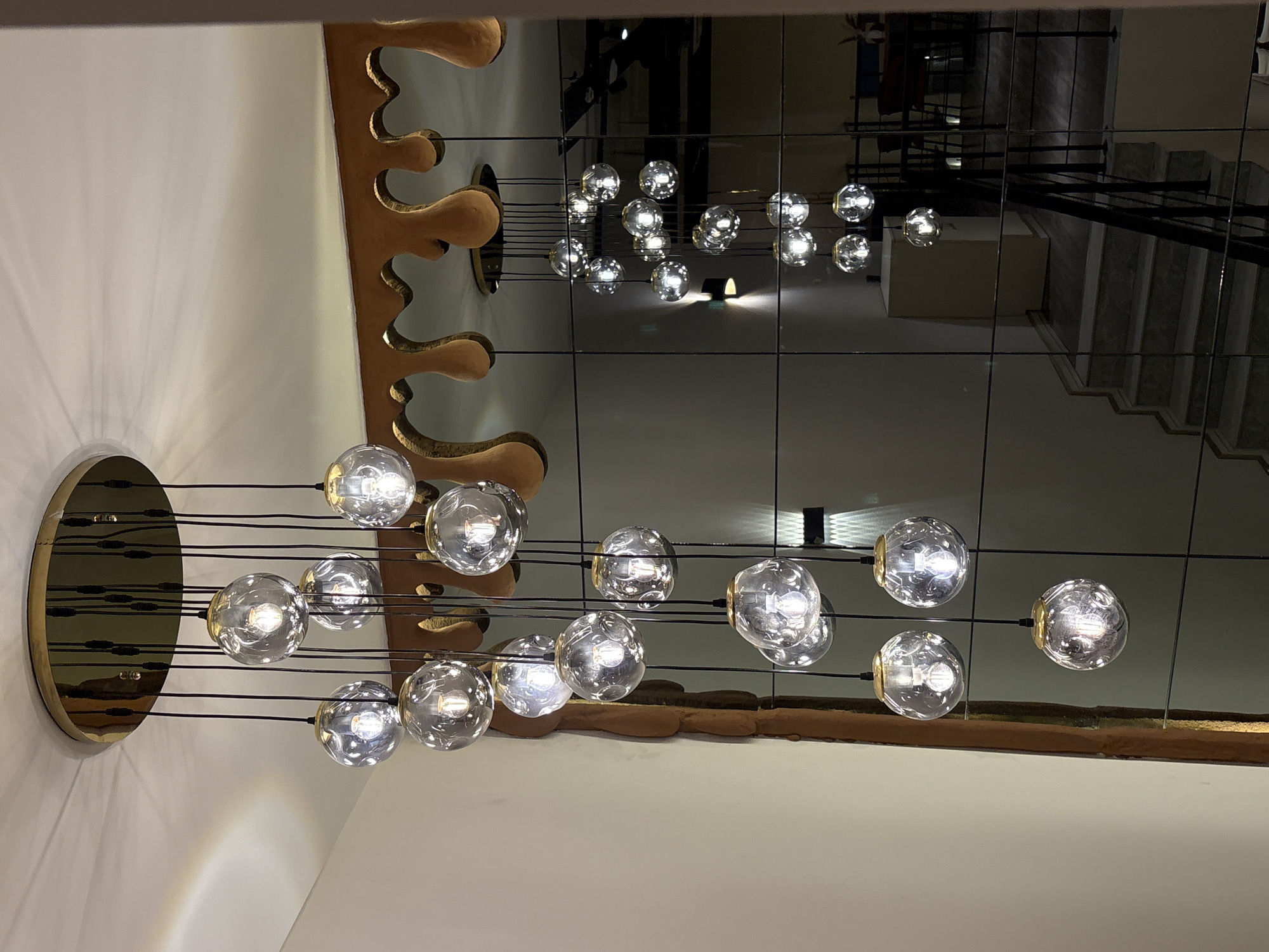 The image shows a modern, multi-tiered chandelier with clear glass globe pendants hanging from a gold-colored ceiling plate. The chandelier is suspended in a well-lit interior space, possibly a residential or commercial building. The pendants are of varying lengths, creating a cascading effect. The background features a large mirrored wall that reflects the chandelier and parts of a staircase, a doorway, and furniture in another room. A decorative, golden, wavy border is visible above the mirrored wall. The overall aesthetic suggests a contemporary and elegant design. The location in Khatay Raion, Azerbaijan, is not discernible from the visual information provided in the image.