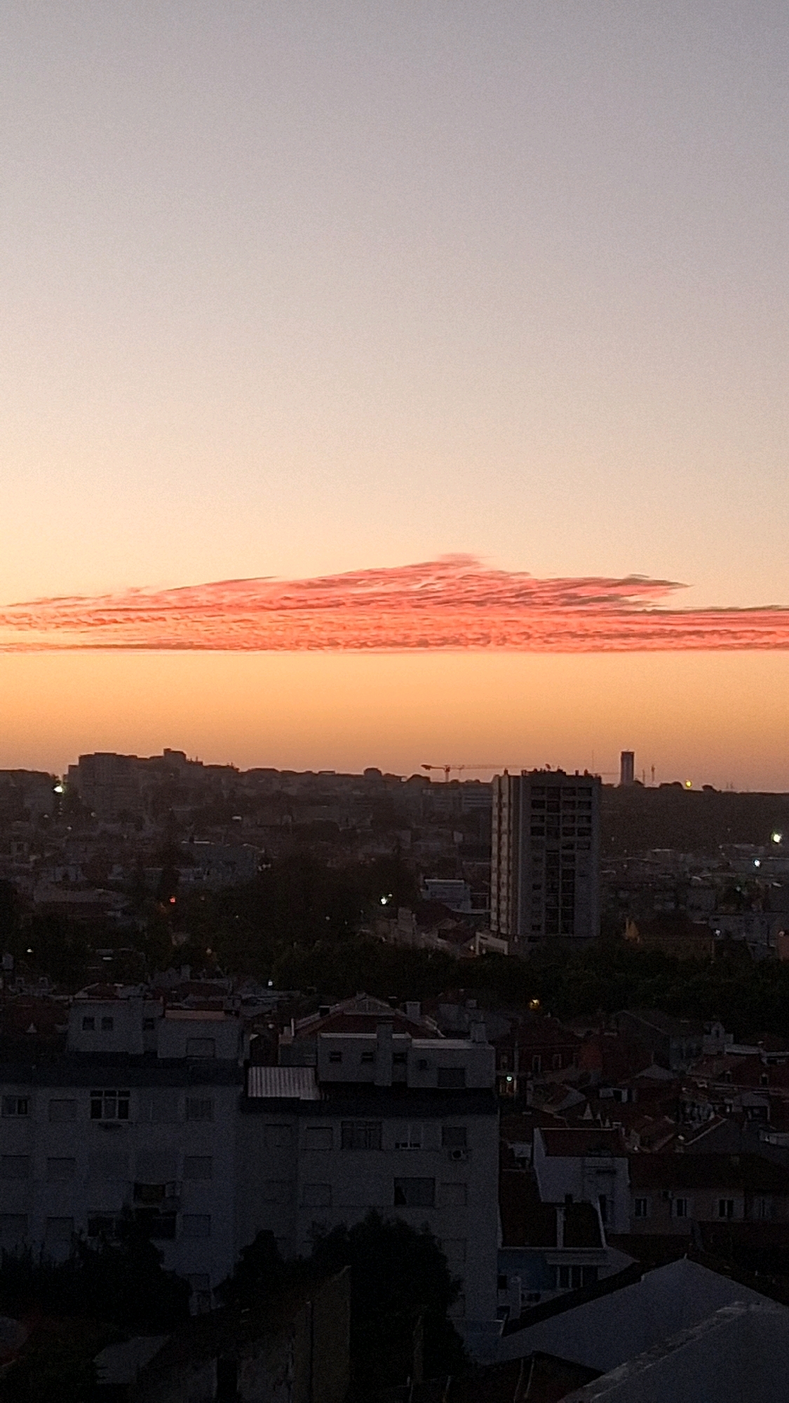 The image shows a panoramic view of a cityscape at dusk. The sky is a gradient of soft orange and grey, with a distinct, long, and layered cloud formation illuminated by the setting sun in a vivid pink and orange hue. Below the sky, the silhouette of a city unfolds, featuring a dense collection of buildings of varying heights and architectural styles. In the foreground and mid-ground, apartment blocks and residential buildings are visible, some with visible windows and balconies, and a few rooftop antennas. The rooftops are predominantly tiled in a reddish-brown color. Further back, taller buildings, including what appears to be a high-rise residential or office building, punctuate the skyline. Construction cranes are also visible in the distance, suggesting ongoing development in the city. The overall impression is a view from a high vantage point overlooking an urban area during twilight. The scene is devoid of any visible people or specific activities, focusing solely on the architectural landscape and the dramatic sky. Without further information, the specific location within Setúbal, Portugal, cannot be identified.