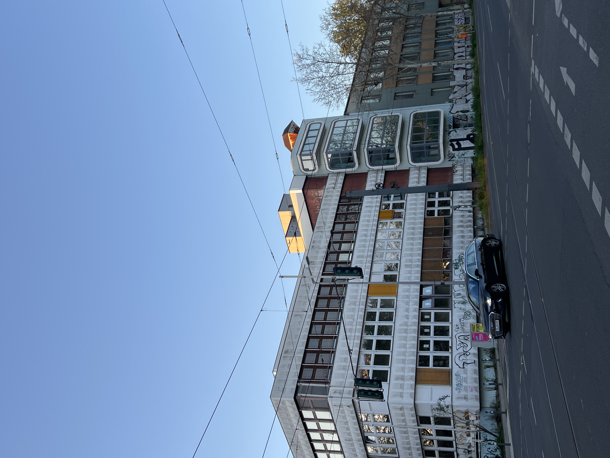 An outdoor urban scene in Berlin, Germany, captured from a low angle, showing a street alongside a prominent building under a clear blue sky. The image is oriented with the sky on the left and the street on the right, as if rotated counter-clockwise.
The left side of the frame is dominated by a multi-story building featuring a textured, light-colored facade with numerous rectangular windows. Some sections of the building are accented with yellow and brown panels. A distinctive architectural element comprises several rounded, protruding window structures or balconies visible at the upper part of this building. Below these, extensive graffiti covers the lower sections of the facade. Above this building, another structure with many smaller, rectangular windows and bare trees in front of it can be seen.
Multiple thin, dark overhead wires stretch diagonally across the sky, likely power lines for trams or trolleybuses, common in Berlin. Two green traffic lights hang from these wires near the main building.
On the right side of the image, a paved road or street is visible, with white dashed lines marking lanes and faint parallel lines that might indicate tram tracks. A dark-colored, possibly black or dark grey, Audi car (an estate/wagon model) is parked along the side of the road. On the car's rear side window, a yellow sign with "STREET FOOD" is visible, and its German license plate reads "B-JJ 4158" (B for Berlin).
The scene is illuminated by bright daylight, suggesting a clear day without clouds, possibly mid-morning or afternoon. No people are visible, and the overall impression is one of an urban streetscape with a mix of architectural styles.