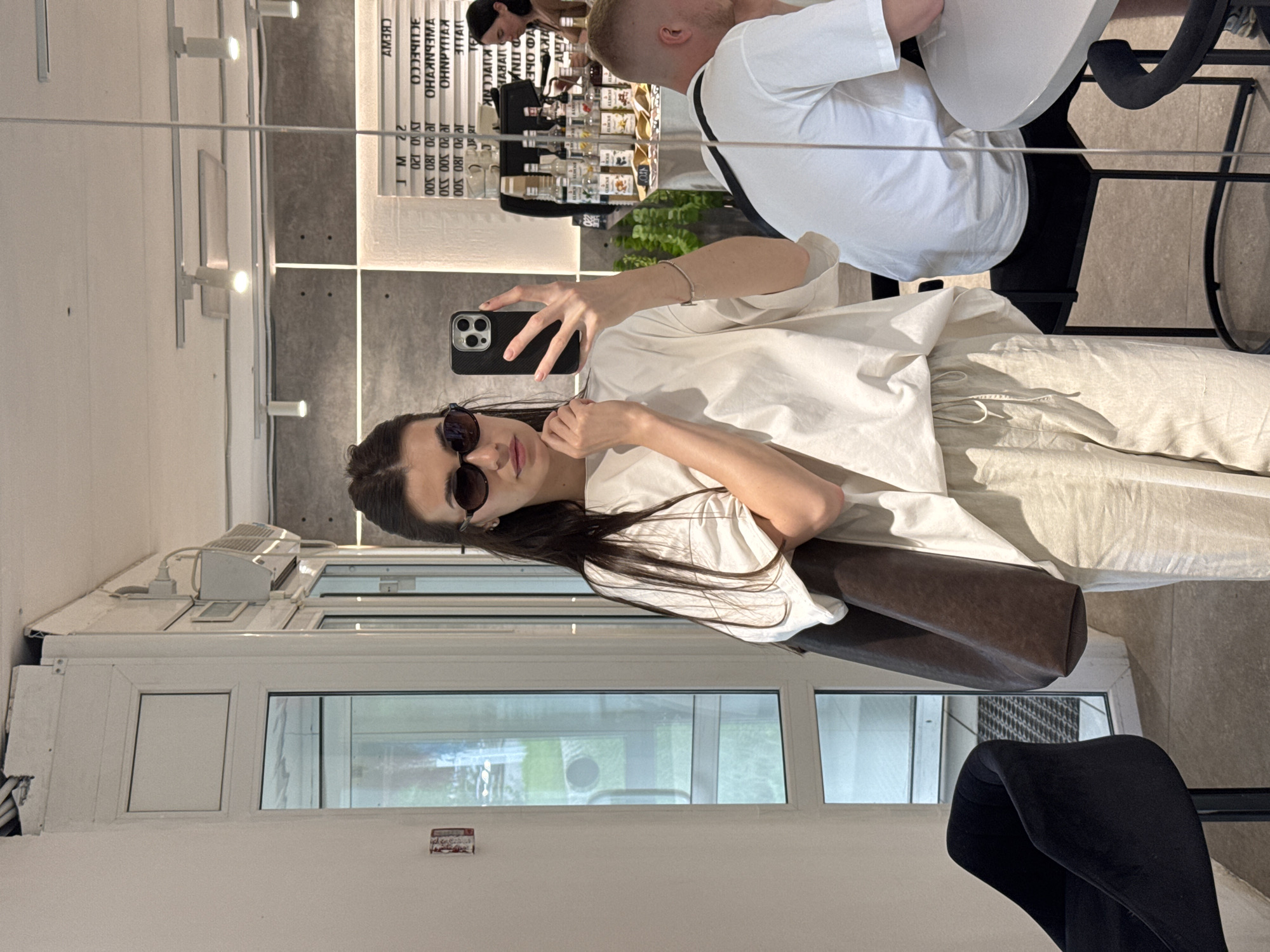 A woman is taking a mirror selfie in a brightly lit, modern cafe. She is wearing an oversized white t-shirt, light-colored linen pants, and dark sunglasses. She is holding a dark brown tote bag in her left arm and a smartphone in her right hand. In the reflection, a man in a white t-shirt is visible sitting at a table, and behind him, a menu board is displayed on the wall. The cafe has a minimalist design with concrete walls and track lighting. The woman's reflection shows a large window on the left and a black velvet chair in the foreground. The location is specified as a cafe in Chaikovsky, Russia.