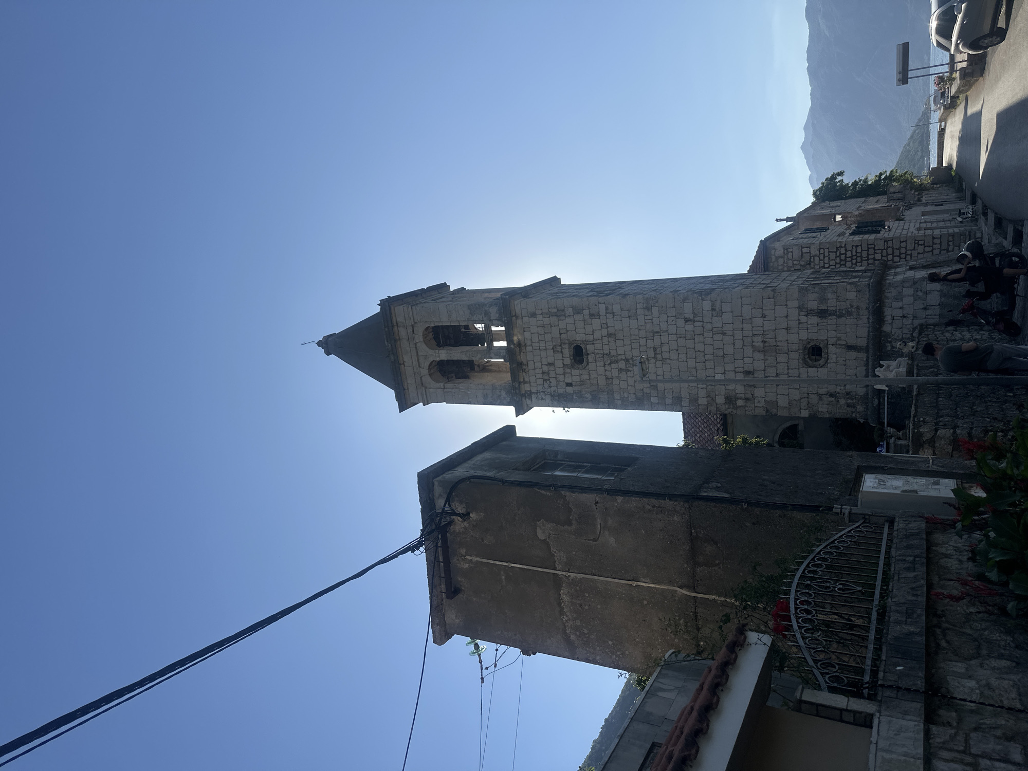 The image depicts a street scene in Prčanj, Montenegro, under a clear, bright blue sky with direct sunlight. A tall, rectangular stone bell tower with a dark, pointed roof topped by a small cross dominates the central right, silhouetted against the sun, which is positioned directly behind it, creating a strong glare and lens flare. Two arched openings for bells are visible near the top of the tower, revealing parts of two bells.
To the immediate left of the bell tower, a weathered, rectangular building with a single visible window on its facade stands. Multiple black electrical wires and cables are attached to its upper section, with a green glass insulator visible. A vertical white pipe runs down its front. In the upper-left, several dark power lines stretch diagonally across the sky.
The scene includes a paved road that curves slightly to the right and descends towards a body of water, visible beyond the buildings on the right. Distant mountains, hazy blue, are seen across the water.
In the lower-left, part of a building with a reddish-brown tiled roof is visible, along with a decorative black metal fence featuring heart-shaped elements, surmounting a stone wall. Red flowers and green foliage grow in the foreground.
Two individuals are on the left side of the road, near the base of the bell tower. One person, wearing a dark t-shirt and light pants, stands facing away from the viewer, looking down the street. Beside them, another person in dark clothing stands next to a dark-colored scooter or moped, which is parked on the side of the road.
Further down the street on the right, a silver or light-colored SUV is partially visible, parked facing away from the viewer. A rectangular street sign or billboard is situated next to it. A stone building with dark green shutters and a wall-mounted light fixture is visible between the bell tower and the road leading to the water. Small white statues or decorative elements are placed on a stone wall at the base of the bell tower. The lighting suggests either morning or late afternoon, with long shadows cast across the road.