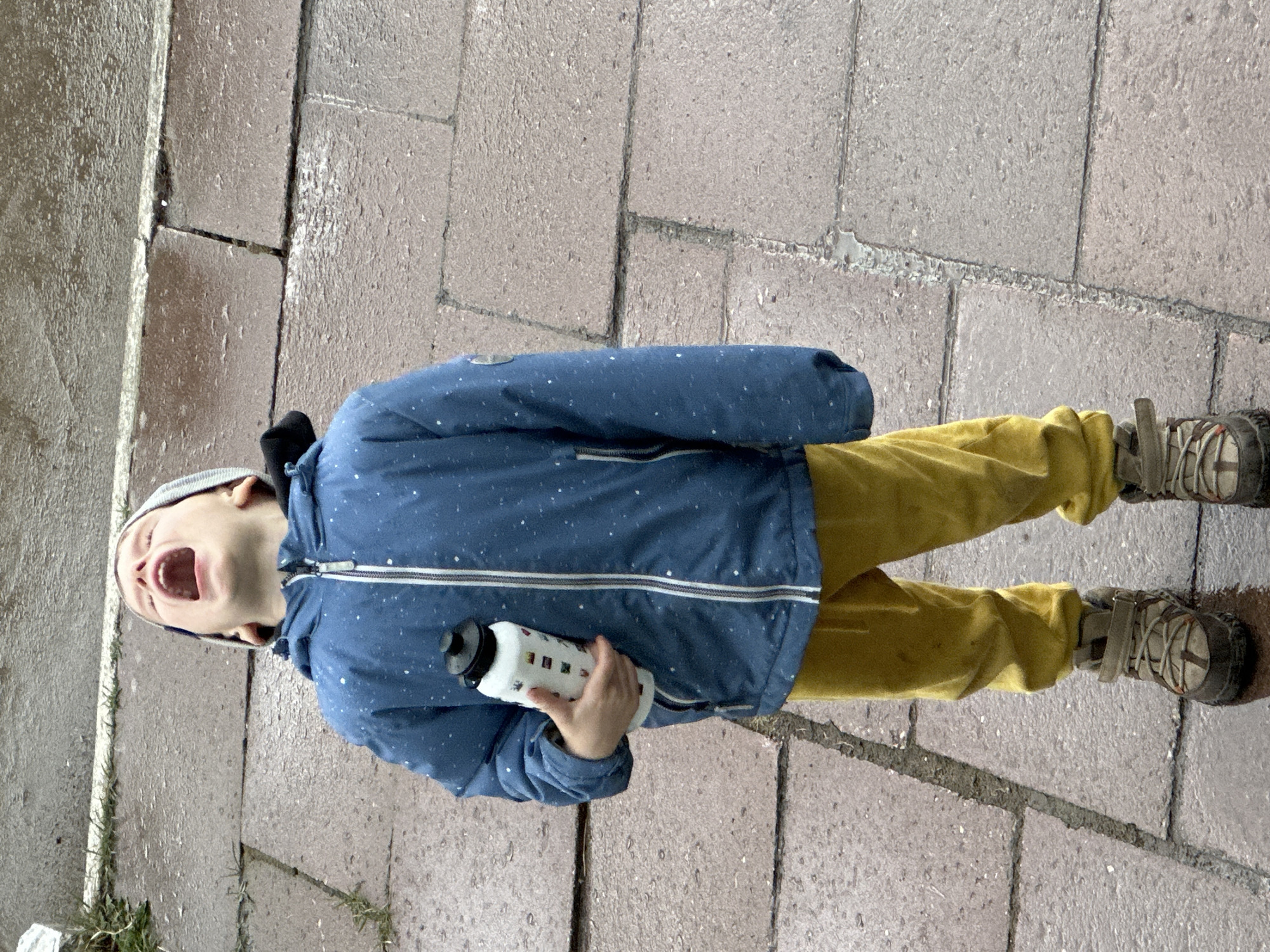 A child stands on a brick-paved surface, possibly a sidewalk or plaza. The child is wearing a blue jacket with white flecks, yellow pants, and hiking-style shoes. The child is also wearing a grey beanie. The child is holding a white water bottle with a black cap and a multicolored square pattern. The child's head is tilted back, and their mouth is open in a scream or yell. The setting appears to be an outdoor area. The location cannot be reliably determined from this image.