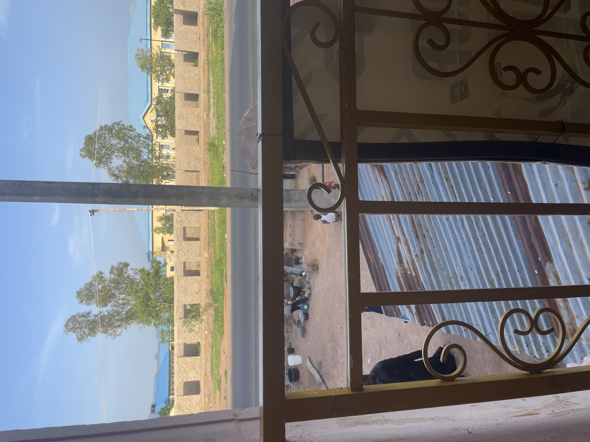 In Jalingo, Nigeria, an outdoor scene unfolds under a bright sky. The foreground features a decorative, golden metal railing, part of a balcony or veranda, framing a view of the surroundings. Below, a corrugated metal roof is visible. Beyond the railing, a dusty courtyard is seen with several people engaged in various activities. Two individuals are tending to cooking fires, with pots and pans scattered around. A young boy sits on a stool, facing away from the camera. Further back, a partially constructed building with visible brickwork and open window frames stands along a paved road. Behind this, a yellow building with a distinctive blue roof is situated, accompanied by lush green trees. The sky above is clear with a few wispy clouds.