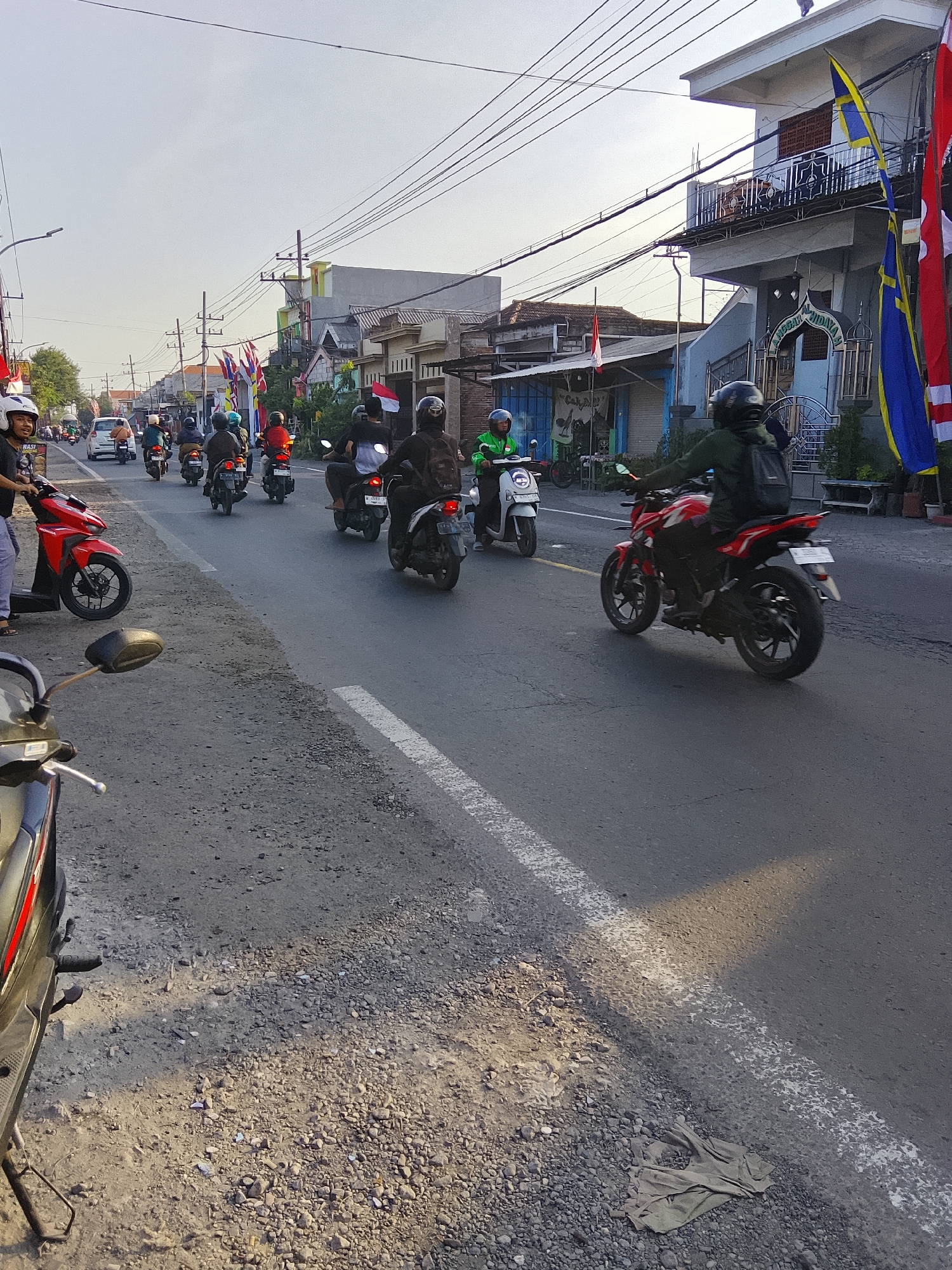The image depicts a street scene in Banjarsari, Indonesia, likely on a sunny day, with a procession of motorcyclists traveling along a paved road. The road is lined with buildings and overhead power lines. Several buildings are visible, some with balconies and architectural details. Flags, including red and white Indonesian flags and blue and yellow flags, are displayed on poles and on some of the buildings.
A group of people are riding motorcycles, mostly moving in the same direction. The motorcyclists are wearing helmets, and some are carrying backpacks. A red motorcycle is prominently featured in the foreground on the right, with its rider wearing a dark jacket and a backpack. Another red motorcycle is parked on the left side of the road. A white scooter is also visible. The road surface is asphalt, with a white line marking the edge of the lane. The area along the side of the road is a mixture of gravel and dirt, with some debris scattered about. The overall impression is of a local street in a populated area.