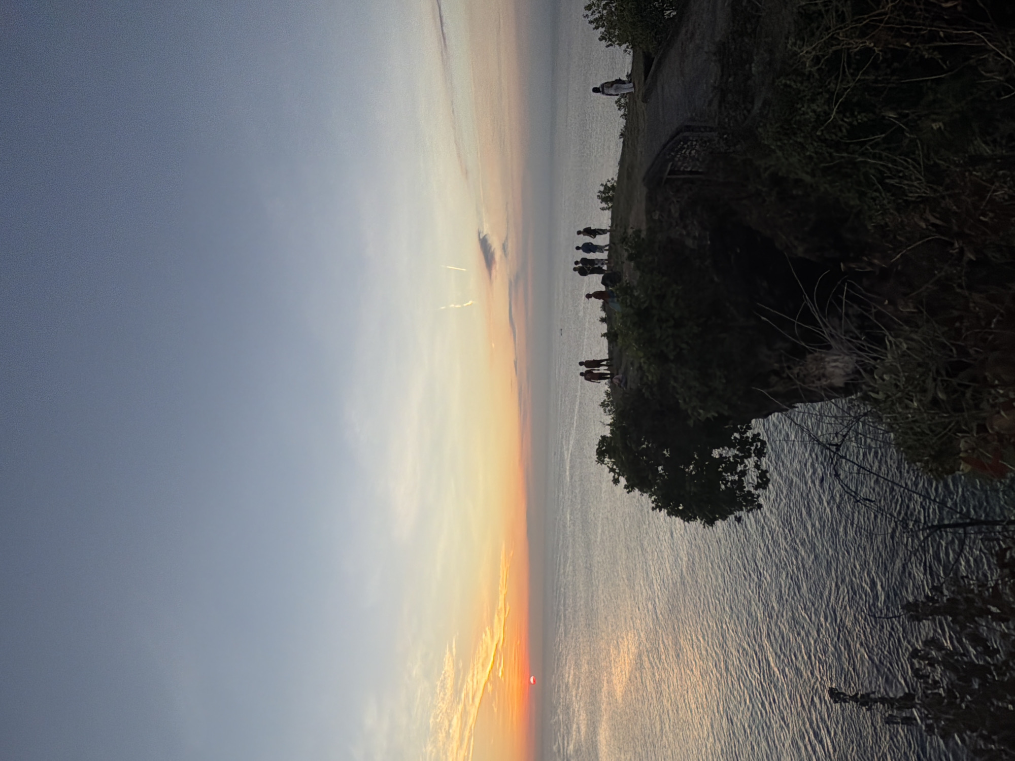 A silhouetted group of people stand on a grassy cliff overlooking a body of water at sunset. The sun, a small red orb, is low on the horizon, casting a warm glow across the sky and water. The sky transitions from a pale blue at the top to shades of orange and pink near the horizon. A few wispy clouds are visible. The cliff is covered in vegetation, with a pathway visible on the right side of the image. The water is calm with gentle ripples. The scene is serene and picturesque, capturing a moment of natural beauty.
