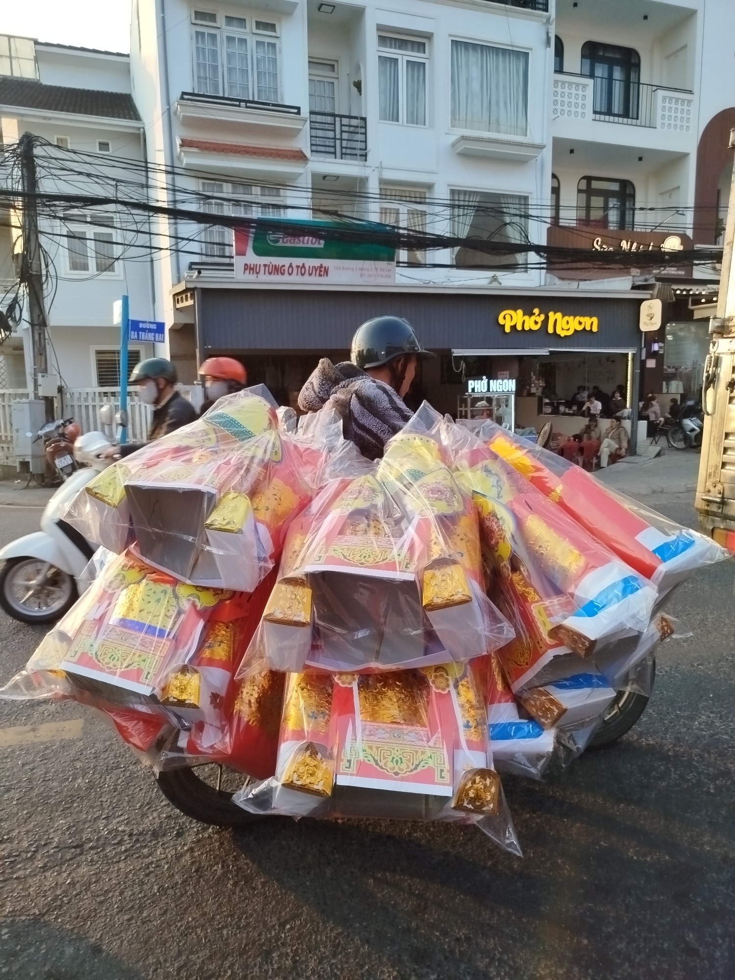 A street scene in Cam Ly - Đà Lạt, Vietnam, features a motorcycle heavily loaded with goods wrapped in plastic, obscuring the rider. The motorcycle is on a paved road. In the background are buildings with businesses; one with a green sign that says “PHỤ TÙNG Ô TÔ UYÊN” and another with the illuminated sign "Phở Ngon." Other people on scooters are on the road. A street sign is barely visible; it reads “BA THÁNG HAI”. Power lines are also visible above.