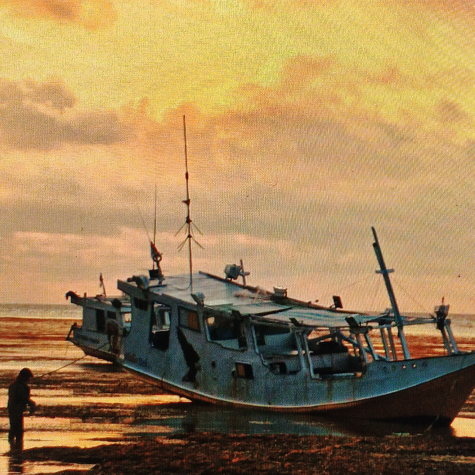 The image depicts a grounded fishing boat on a shallow shoreline, likely during low tide, with a person standing near the bow. The boat is a prominent, faded blue structure with visible signs of wear and tear. It has multiple decks, railings, and masts, suggesting it's a working vessel. The person, silhouetted and facing away from the camera, appears to be inspecting or interacting with the boat. The environment is a beach with wet sand reflecting the warm hues of the sky, which is characterized by a gradient of yellow and orange, indicative of either sunrise or sunset. The overall impression is one of stillness and a moment captured at the edge of the water. The scene is consistent with the coastal areas of Ende, Indonesia, where such fishing boats are common and low tide conditions frequently expose shorelines.