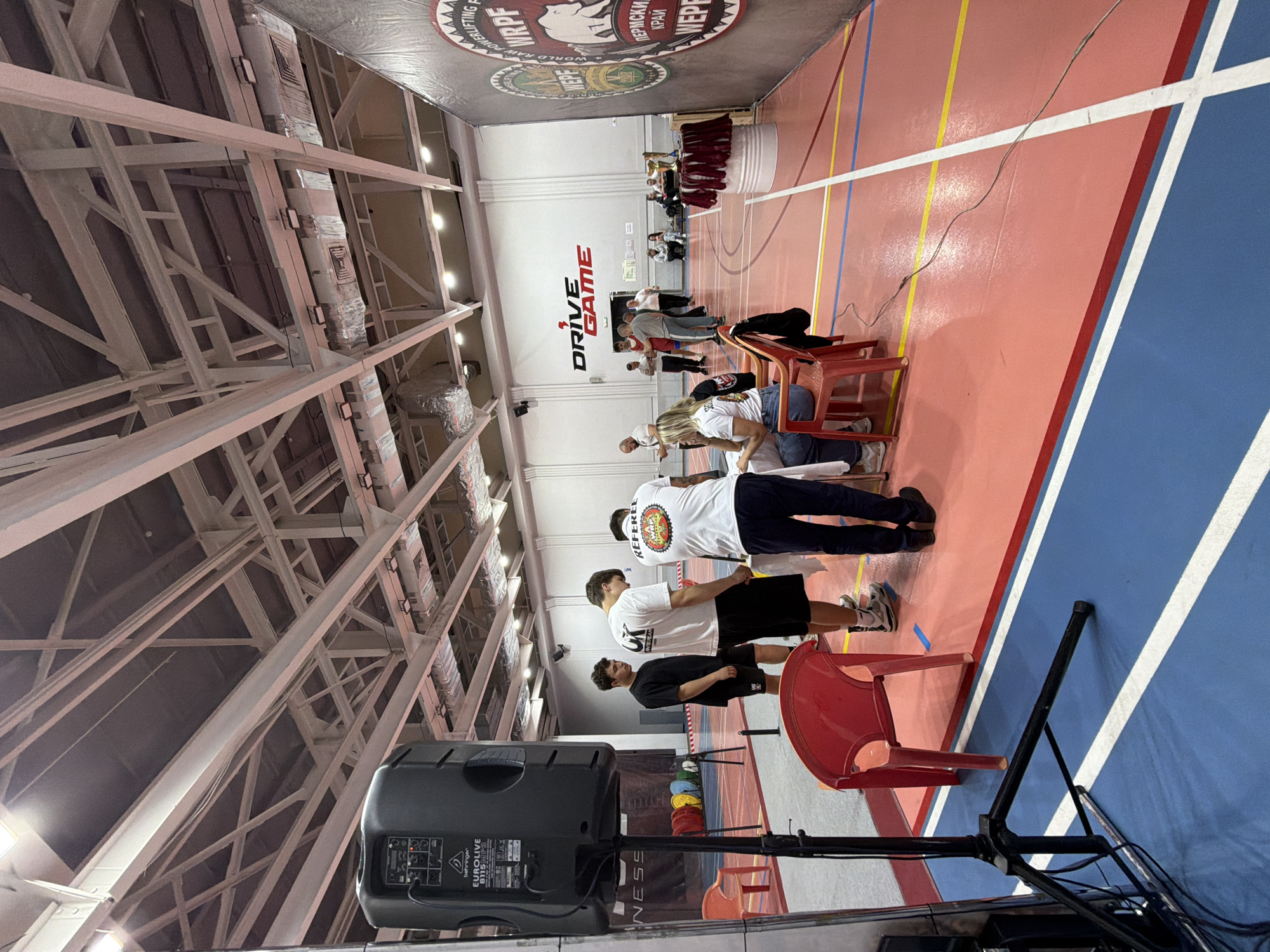 The image shows the interior of what appears to be a sports facility or gymnasium. The space is painted with red, yellow, white, and blue lines, likely marking playing areas. In the background, there is a large banner with the text "WORLD RAW POWERLIFTING FEDERATION WRPE" and the text "ПЕРМСКИЙ КРАЙ" which translates to "Perm Krai", indicating the location is within Perm Krai, Russia.
Several people are standing or sitting on chairs, seemingly observing or participating in an event. One man is wearing a white shirt with a logo on the back. A black speaker is visible in the foreground. The building has a high ceiling with exposed steel beams and ductwork.