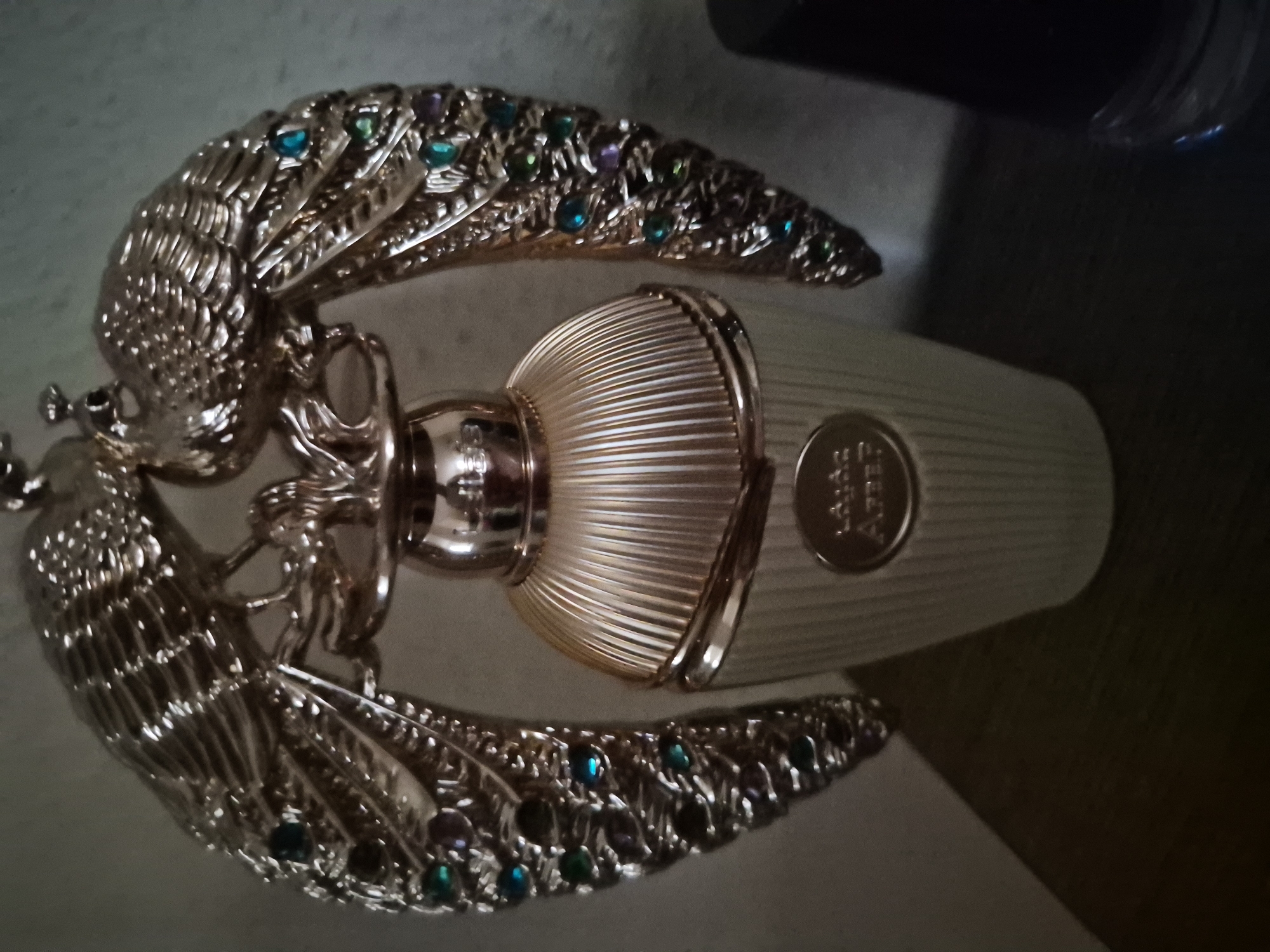 An ornate gold perfume bottle with a peacock motif sits on a dark surface. The bottle's base is ribbed and pale gold, with a circular emblem in the center bearing an inscription. Above the bottle, two gold peacocks with spread wings and jeweled accents form a decorative cap. The peacocks' wings are adorned with colorful gemstones, including blue, green, and purple. The background is a plain, light-colored wall. The lighting casts a shadow from the bottle onto the surface.