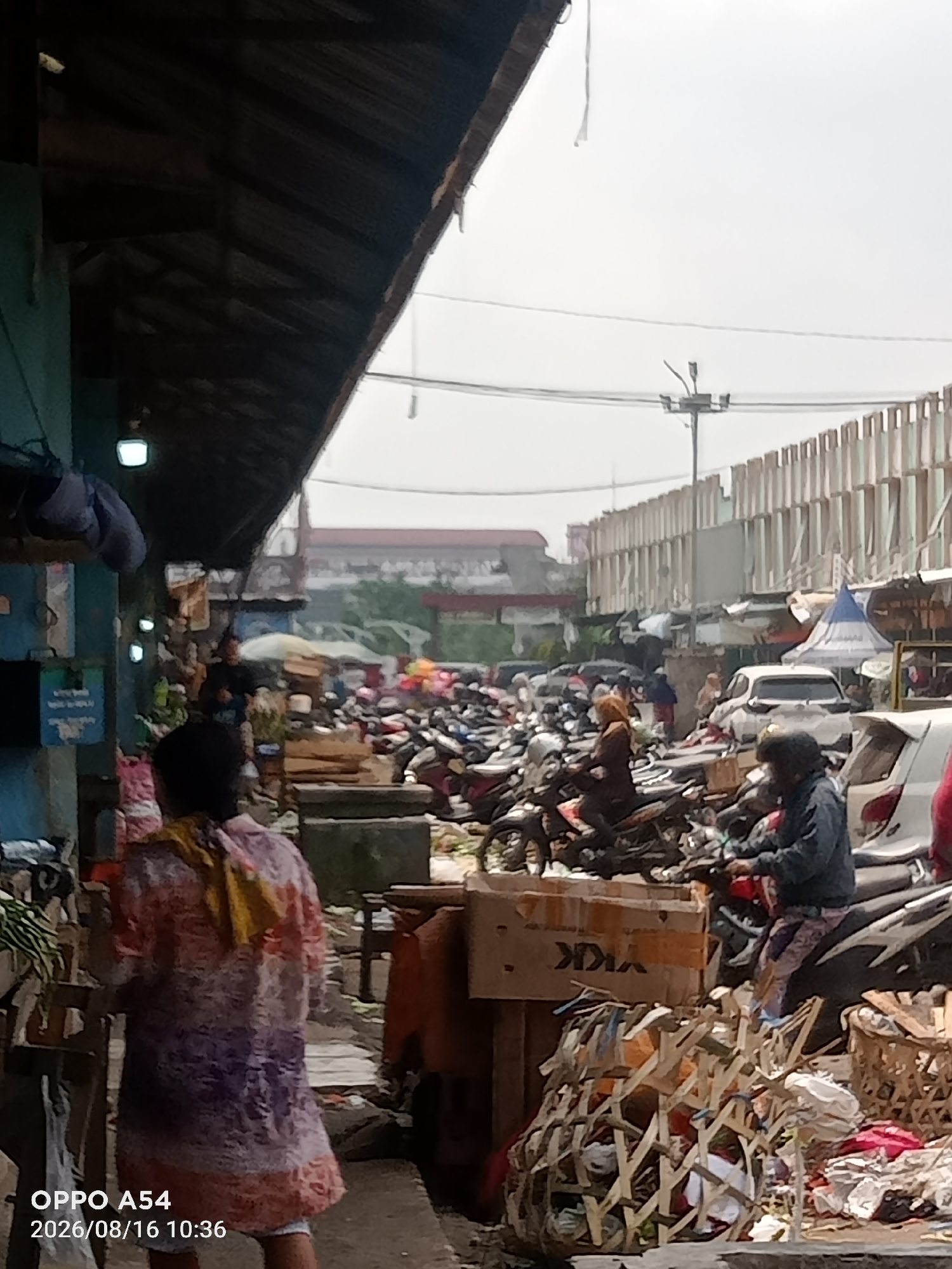 This is a medium-shot, horizontal, outdoor photograph taken at a market in Jambi City, Indonesia, on August 16, 2026, at 10:36 AM. The image captures a bustling marketplace scene with numerous motorcycles parked and people moving around. In the foreground, a person wearing a colorful patterned garment with purple, pink, and orange hues is facing away from the camera. To their right, a person in a dark jacket and patterned pants is seated on a motorcycle. Various market stalls are visible, with wooden structures and some covered areas providing shade. In the mid-ground, a dense collection of motorcycles dominates the scene, with some individuals pushing or maneuvering them. Behind the motorcycles, market tents and structures are visible, along with more parked vehicles. The background features a long, multi-story building with vertical paneling and a bright, overcast sky. The overall atmosphere suggests a busy commercial area with a significant amount of pedestrian and vehicular traffic. The presence of the "OPPO A54" watermark in the lower left corner indicates that the photograph was taken with an Oppo A54 smartphone.