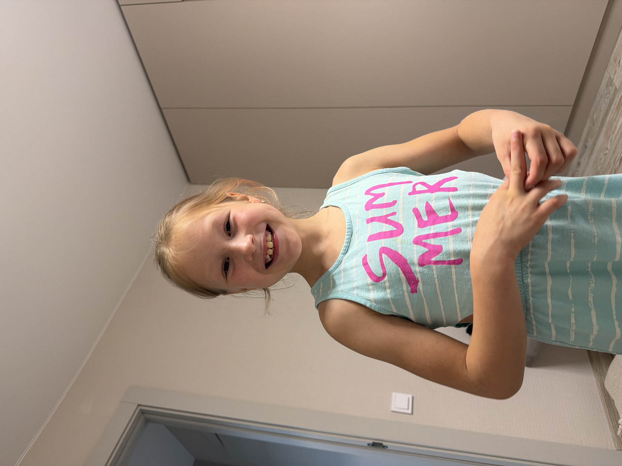 The image features a young girl with blonde hair, smiling broadly and looking at the camera. She is wearing a light blue and white striped tank top with the word "SUMMER" printed in large pink letters. Her arms are raised, and her hands are positioned near her chest, with her fingers interlaced. The girl's expression conveys happiness and playfulness.
The setting appears to be indoors, possibly in a home, with neutral-colored walls and cabinetry in the background. A doorway with a light-colored frame is visible at the bottom of the image. The lighting suggests daytime. The image is oriented sideways, with the girl appearing to be upside down relative to a standard upright orientation.
The visible text on the girl's shirt reads "SUMMER". The overall impression is a candid and joyful moment captured with a child.