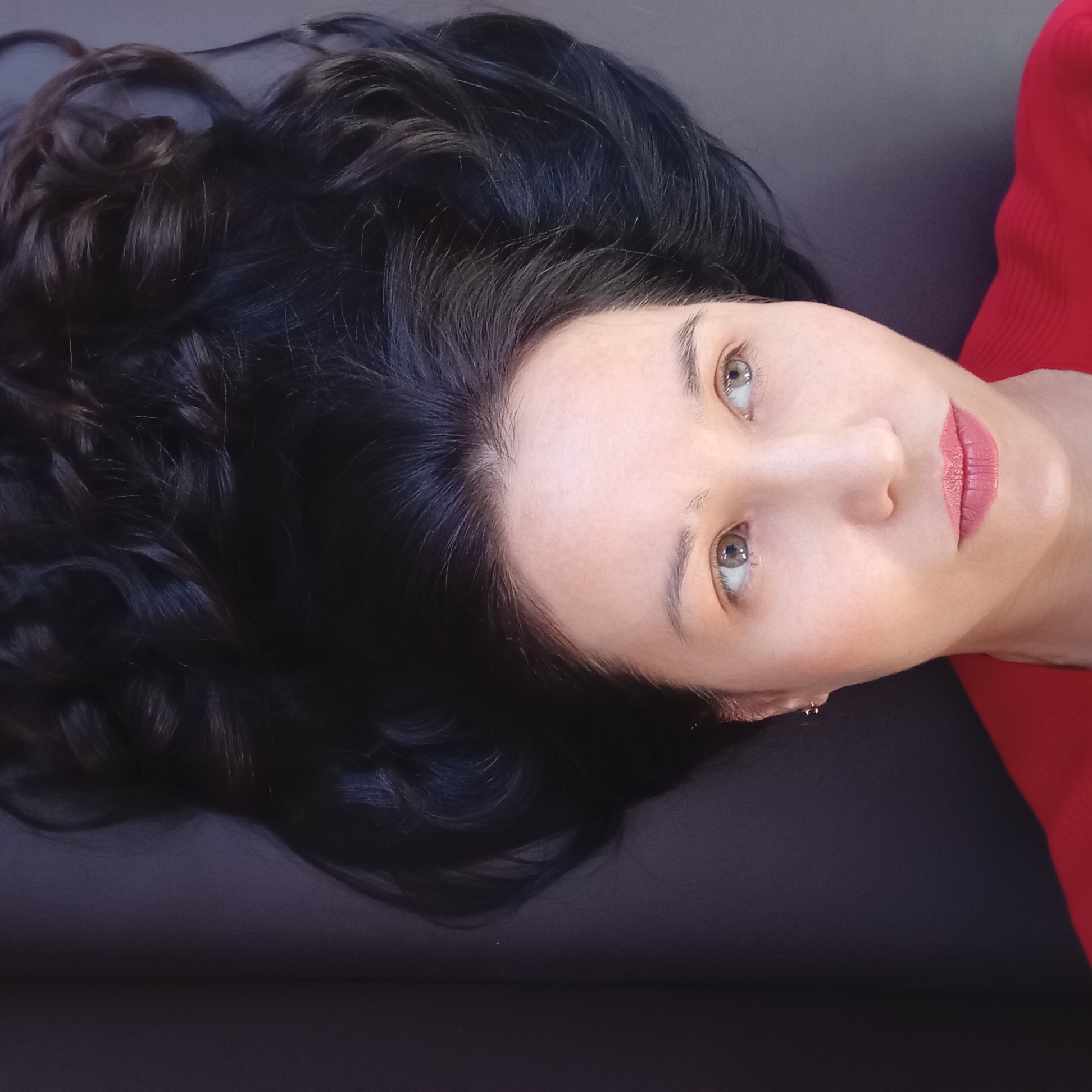 A woman with dark, wavy hair is lying down on a dark surface. Her head is positioned at the top of the frame, with her hair spread around her. Her face is visible, and she appears to be looking upwards. She has light-colored eyes and is wearing lipstick. A red garment is visible on the right side of the image. The location is not discernible from the given image.