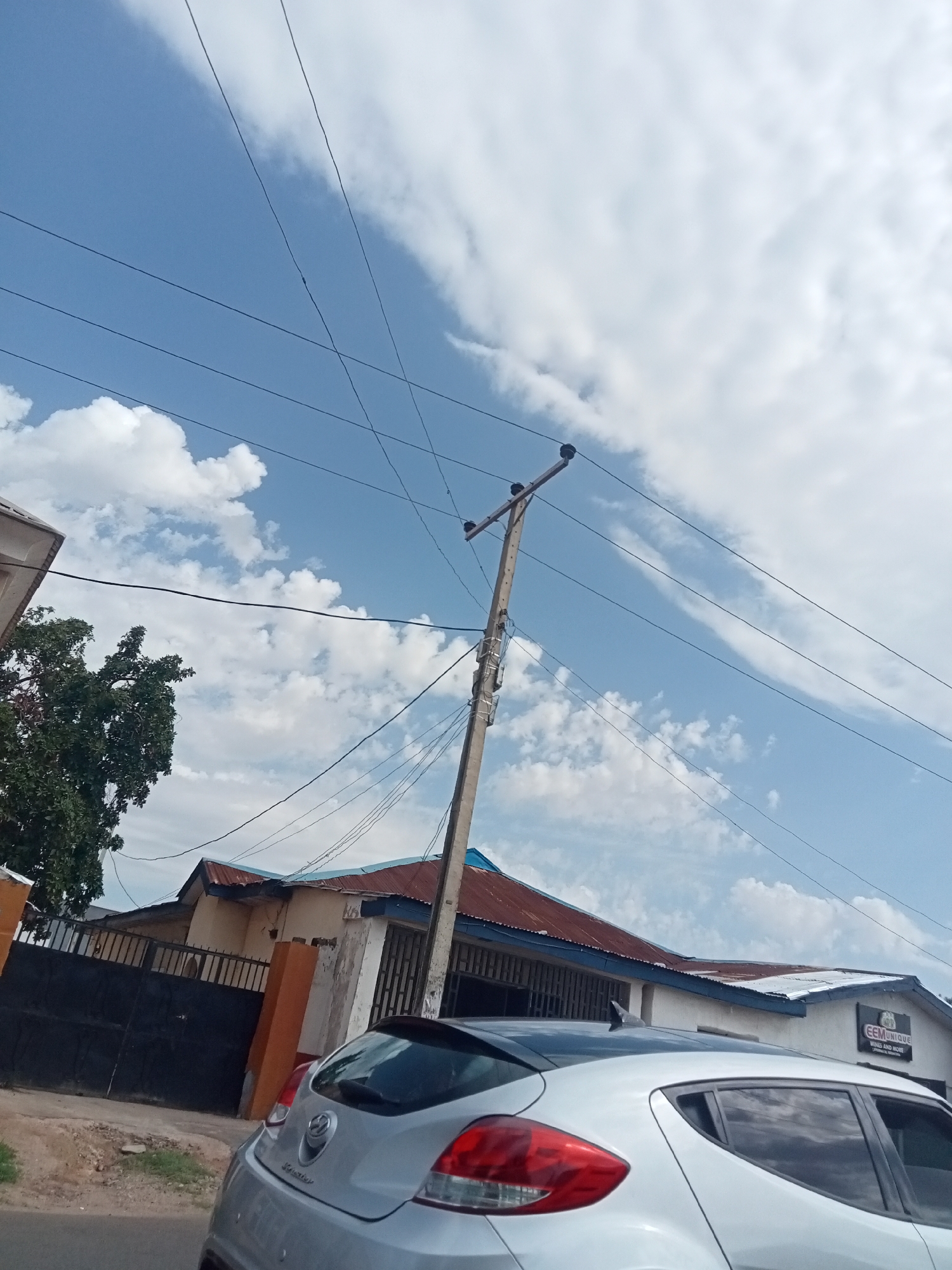 The image shows a street scene in Jos, Nigeria. A silver Hyundai Veloster is parked on the right side of the frame, partially obscuring the view of a building with a sign reading "EEMUNIQUE WINES AND MORE". The building has a white facade, a corrugated metal roof, and a blue trim. A black metal gate and a house with orange pillars are visible behind the Veloster. Overhead, an electric utility pole with multiple wires spans across the sky. The sky is blue with scattered white clouds. The environment suggests a residential or commercial area. No people are directly visible.