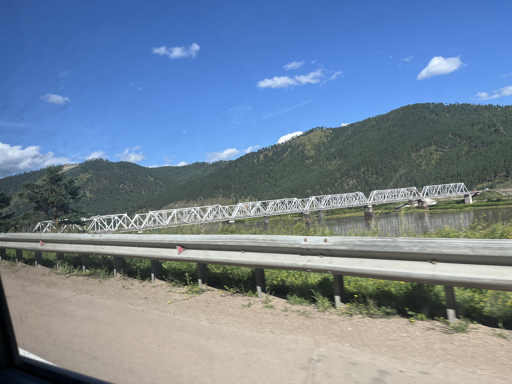 A wide-angle, eye-level shot shows a long, white metal truss bridge spanning a wide, calm river. The bridge is supported by multiple concrete piers and has a distinctive lattice structure. On the far bank of the river, a dense, green forest covers the slopes of rolling hills that recede into the distance under a clear, bright blue sky with scattered white clouds. In the foreground, a grey metal guardrail runs along the edge of a dirt road, with sparse green vegetation and some yellow wildflowers growing beside it. The overall scene is one of natural beauty and infrastructure, likely captured from a moving vehicle.