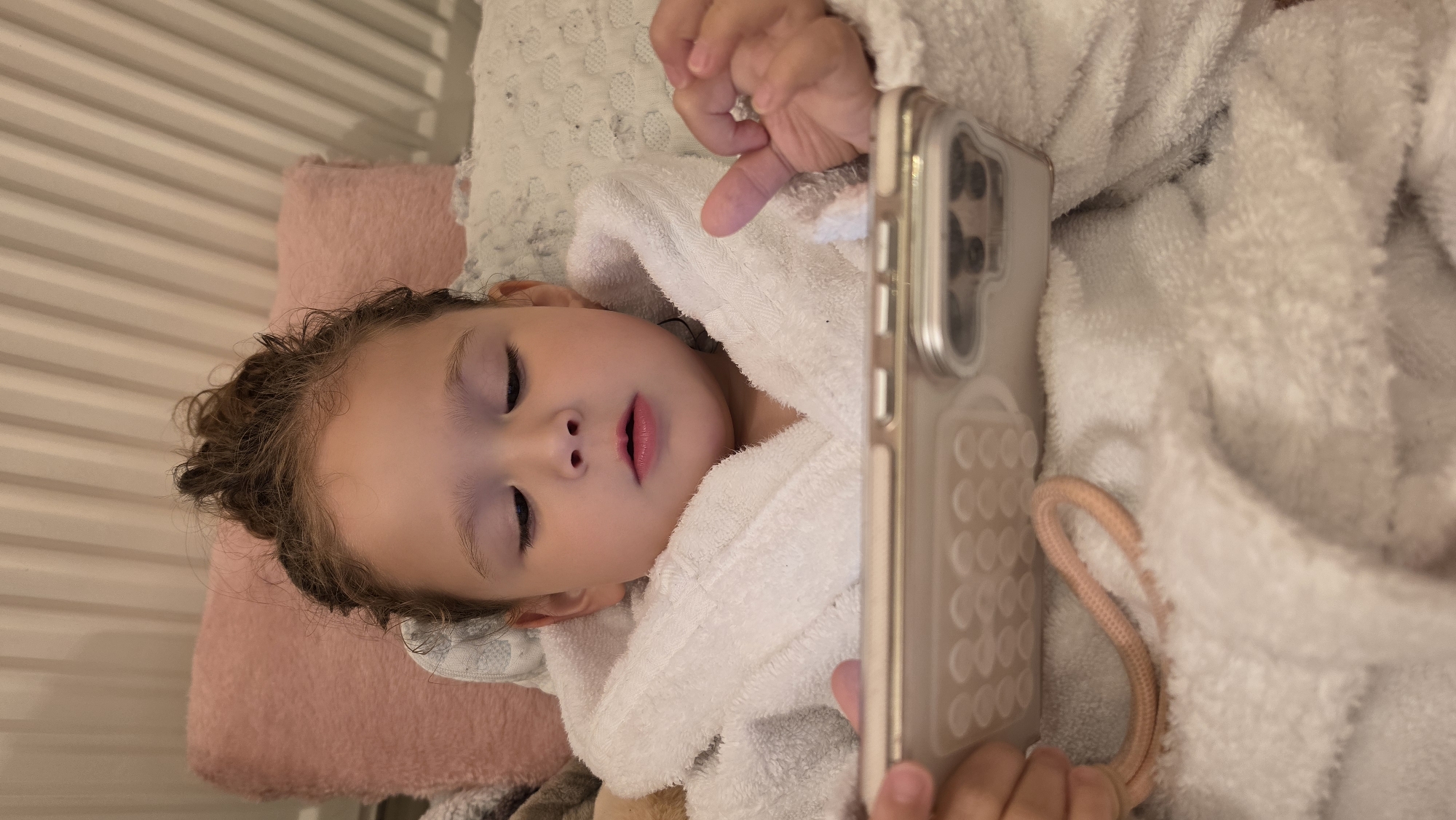 A young child is lying down, possibly in bed, wearing a white hooded bathrobe. The child is looking at a smartphone, which is held up in front of them. The child is lying on a pink pillow, and there is another pillow visible behind it. The background shows striped walls. The location is indeterminate and not clearly identifiable as London, United Kingdom.