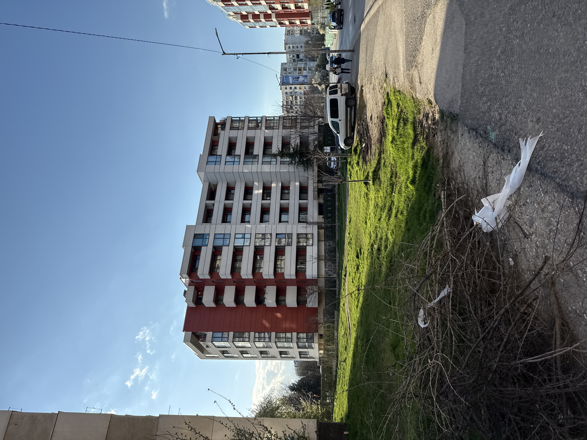 The scene depicts a building in Sofia, Bulgaria, with a red and white facade. A street runs along the side of the building, lined with grass and debris, including a white plastic bag tangled in branches. A white van and two pedestrians are visible further down the street. Other buildings are in the background. The sky is blue with scattered clouds.