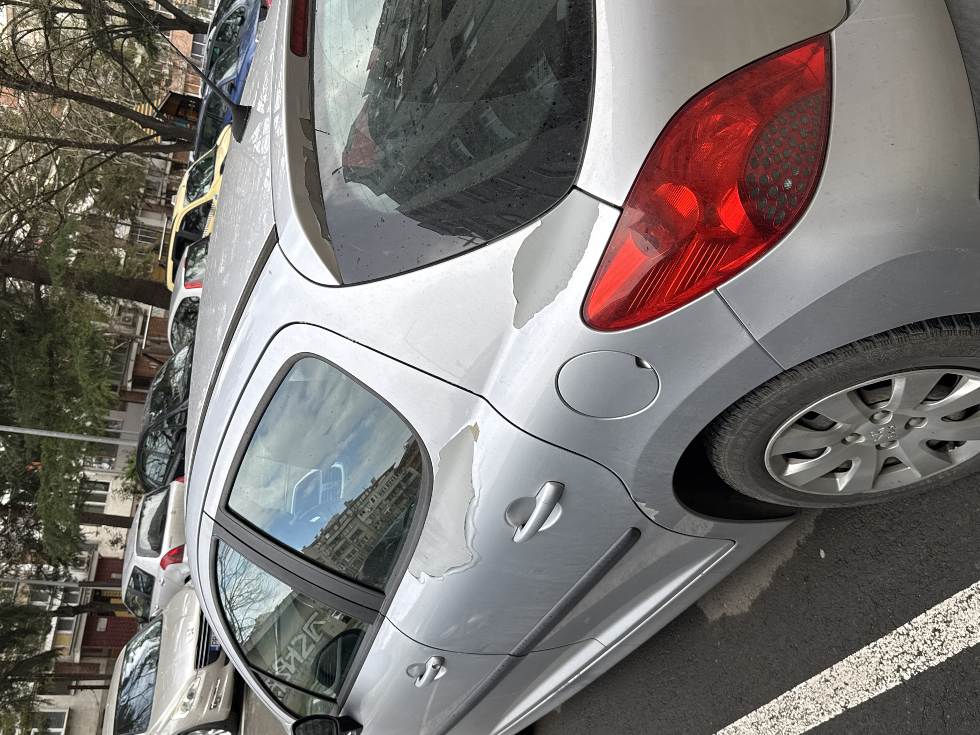The image depicts a silver Peugeot hatchback parked in a row of cars on a street in Burgas, Bulgaria. The car's paint is peeling near the rear window and the area around the fuel door. The car is parked on asphalt next to a white painted line. Other cars of various colors are parked in a row behind the Peugeot. Buildings and bare trees are visible in the background. Reflections of buildings are visible in the car's windows.
