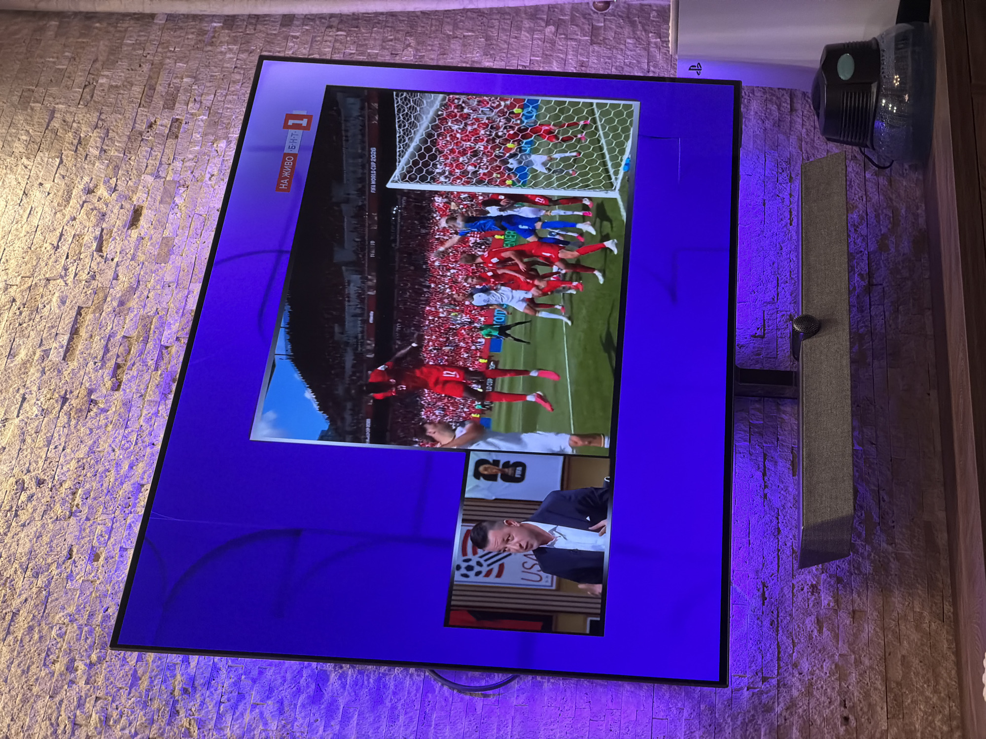 The image shows a television screen displaying a soccer match, likely the FIFA World Cup 2020. The screen is mounted on a wall covered with textured stone tiles, illuminated by purple LED lights. A soundbar is mounted below the television. On the right side of the television, a white PlayStation console is visible. In the foreground, a wooden surface holds a humidifying device. The television screen itself is split into two parts. The larger part shows a live soccer game with players in red and white uniforms. A smaller inset screen on the left shows a man in a suit speaking, with a graphic displaying "USA" and a soccer ball behind him. The broadcast seems to be in Russian, as indicated by the "НА ЖИВО" text on the screen. This scene is likely in a domestic setting in Sofia, Bulgaria, given the presence of the PlayStation console and the broadcast language.
