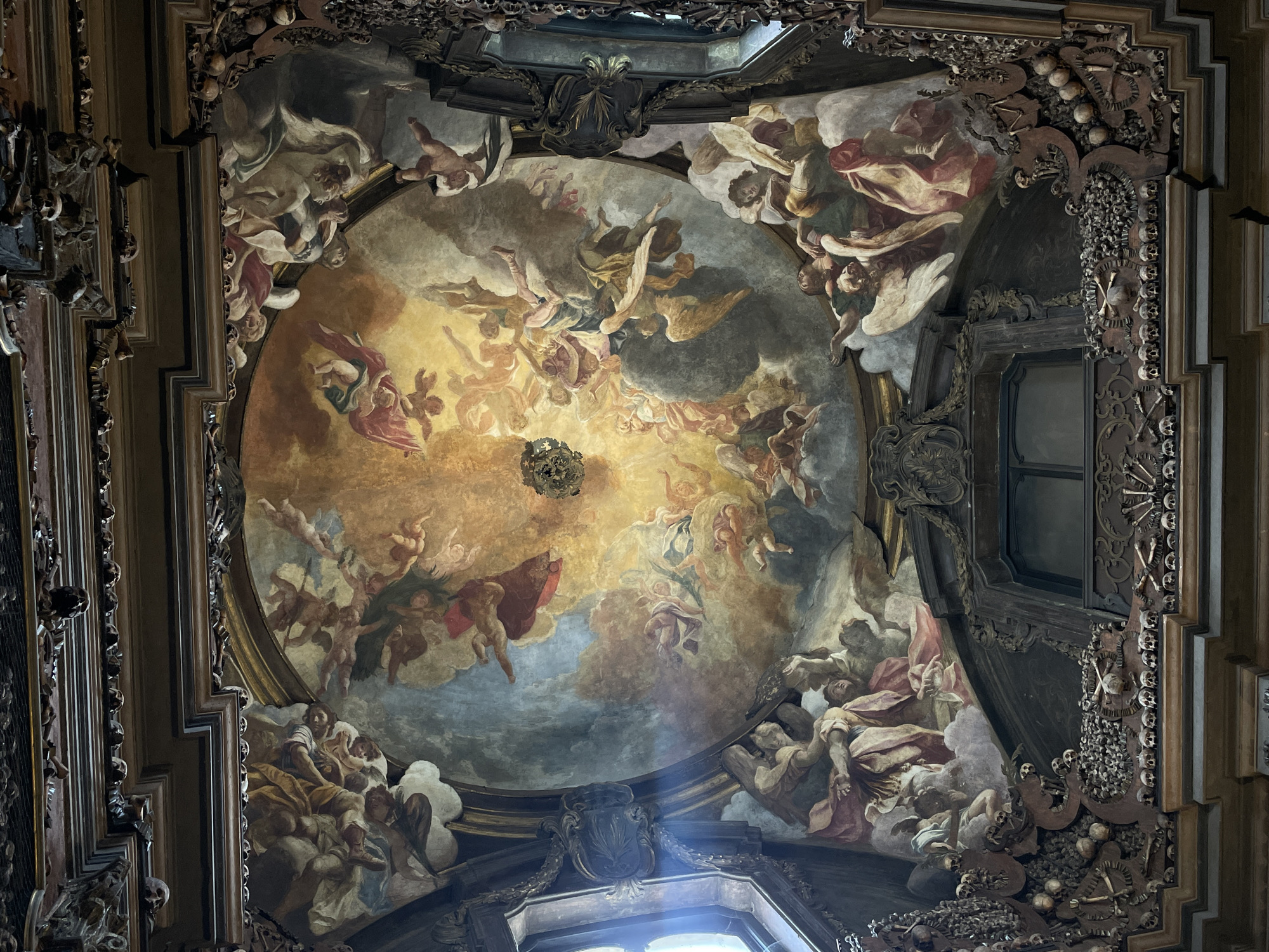 The image captures a view of the ceiling inside the San Bernardino alle Ossa church in Milan, Italy. The ceiling features a painted fresco depicting numerous figures of angels and cherubs against a background of clouds and golden light emanating from the center. The scene is framed by a heavily ornamented border of wood and plaster, which includes skulls and bone motifs. A window is visible on the right, and beams of light highlight architectural details. The overall style is baroque.